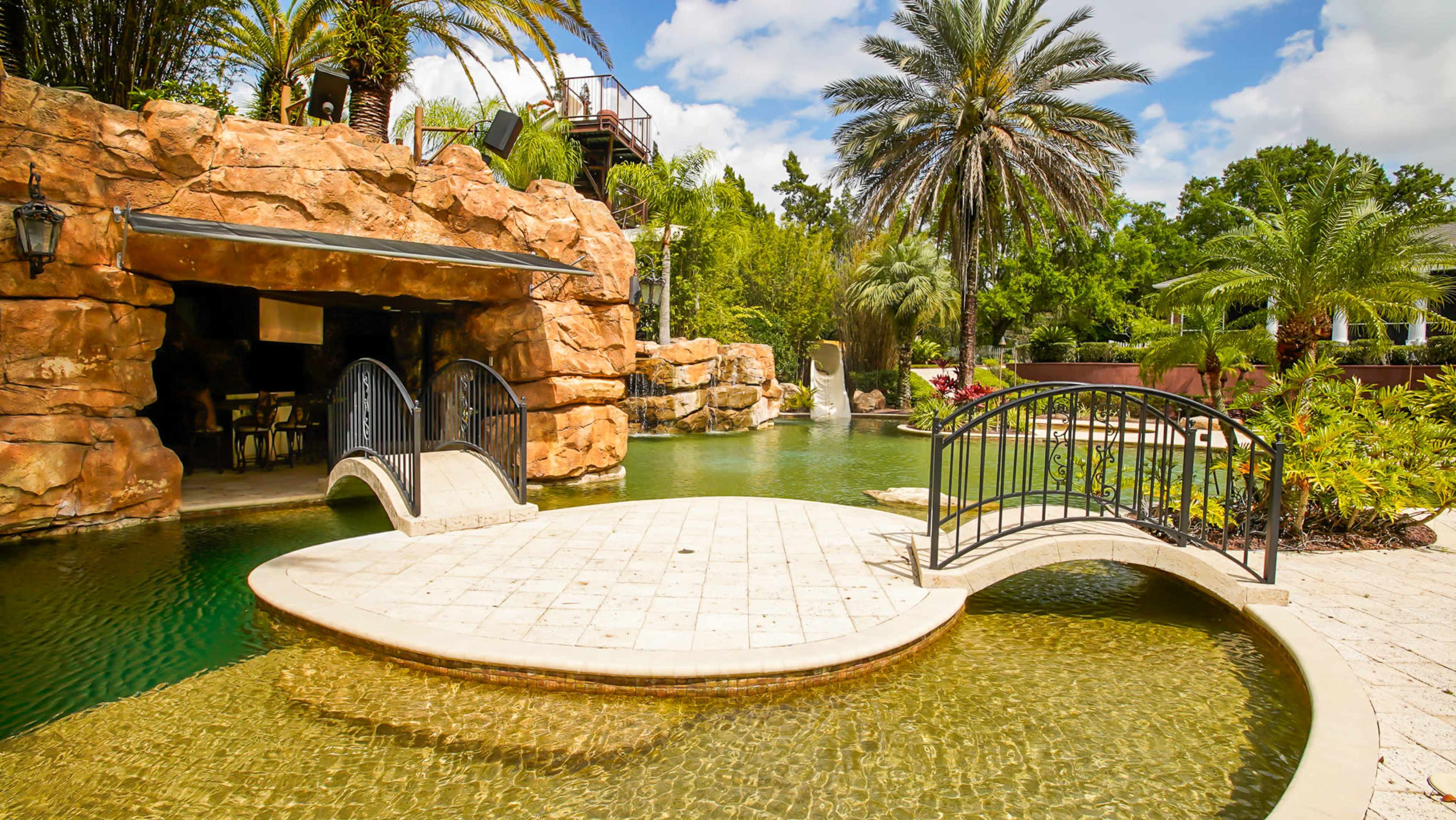 A landscaped outdoor space featuring a circular stone platform connected by a small bridge to a rocky cave-like structure, surrounded by palm trees and water.