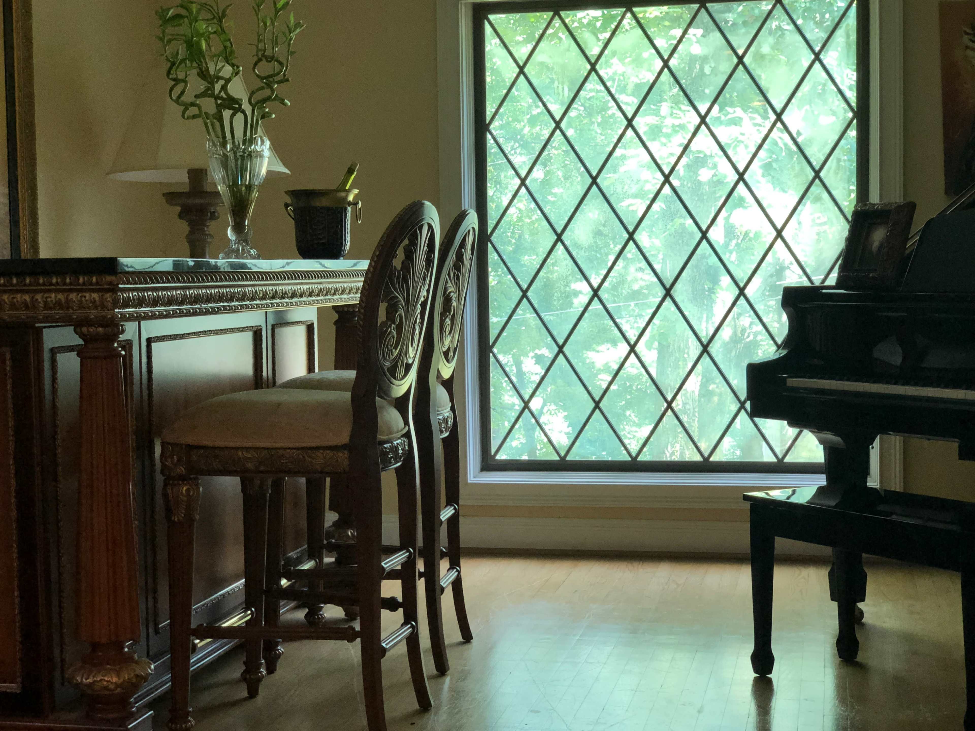 A wooden bar area with three high stools, a lamp, and a large diamond-patterned window letting in light, alongside a black piano.