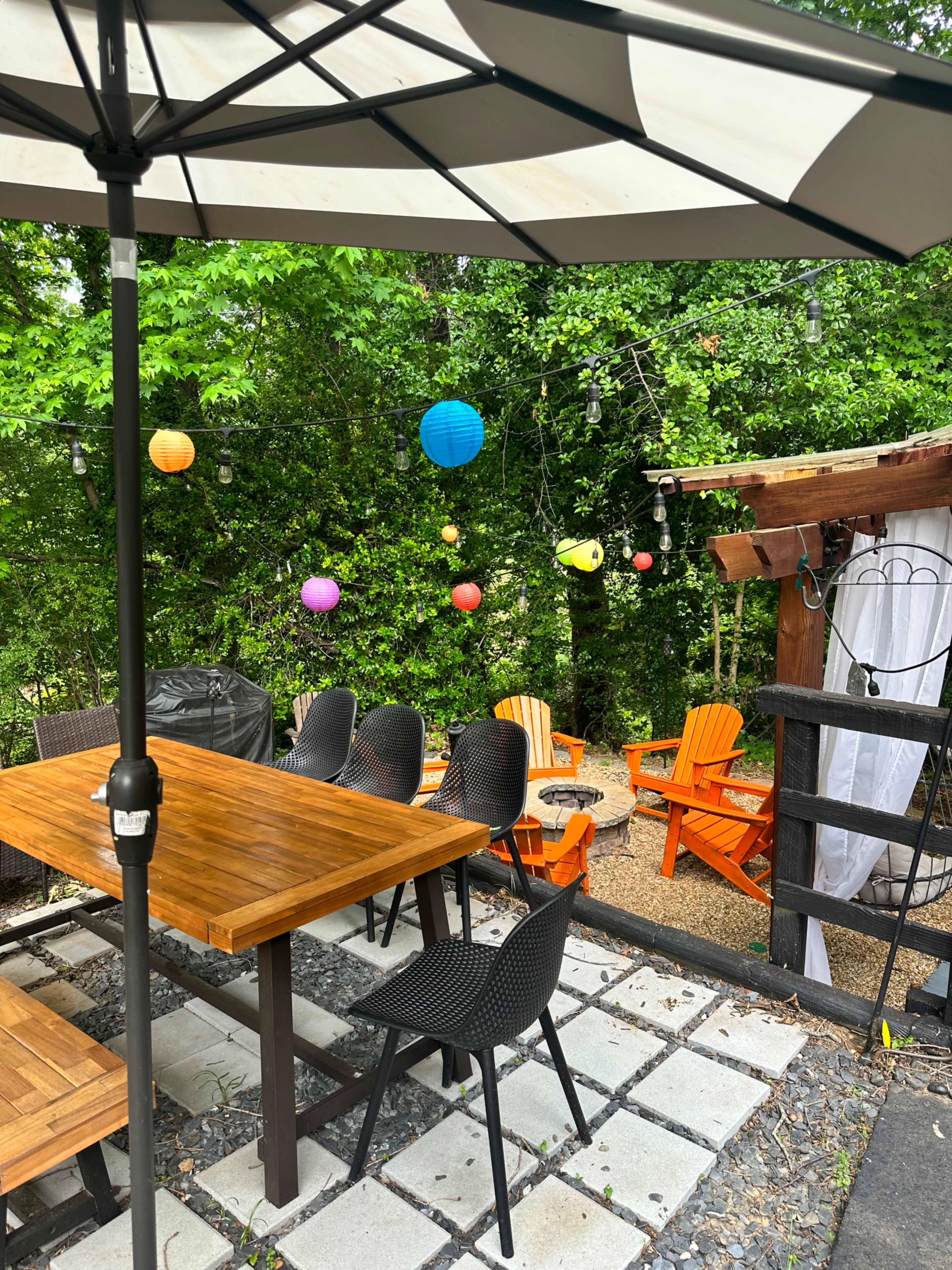 A patio area features a wooden table and chairs, surrounded by colorful lanterns hanging from trees.