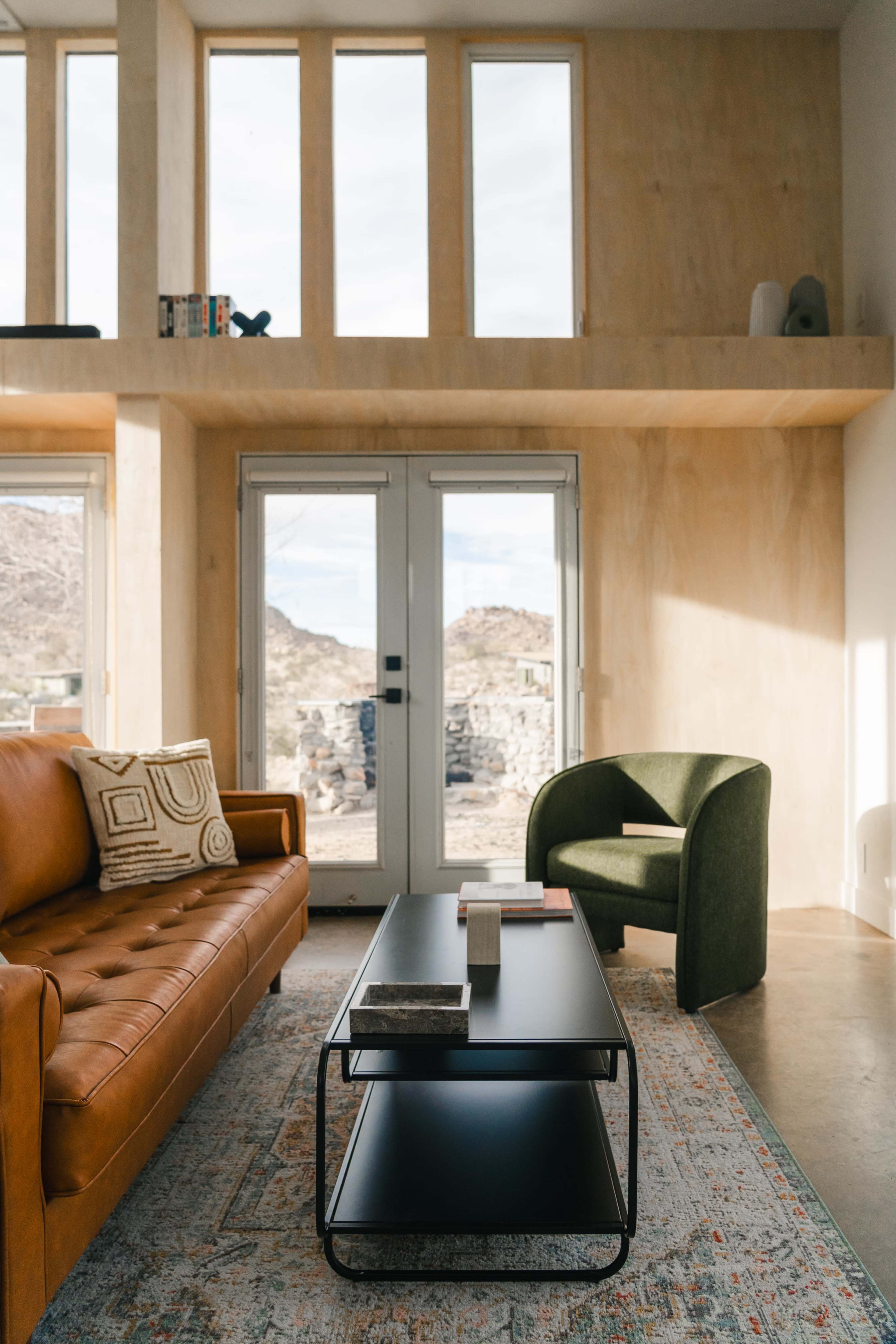 The image shows a modern living room with a brown leather sofa, a black coffee table, and a green upholstered chair, featuring large windows that provide a view of the outdoors.