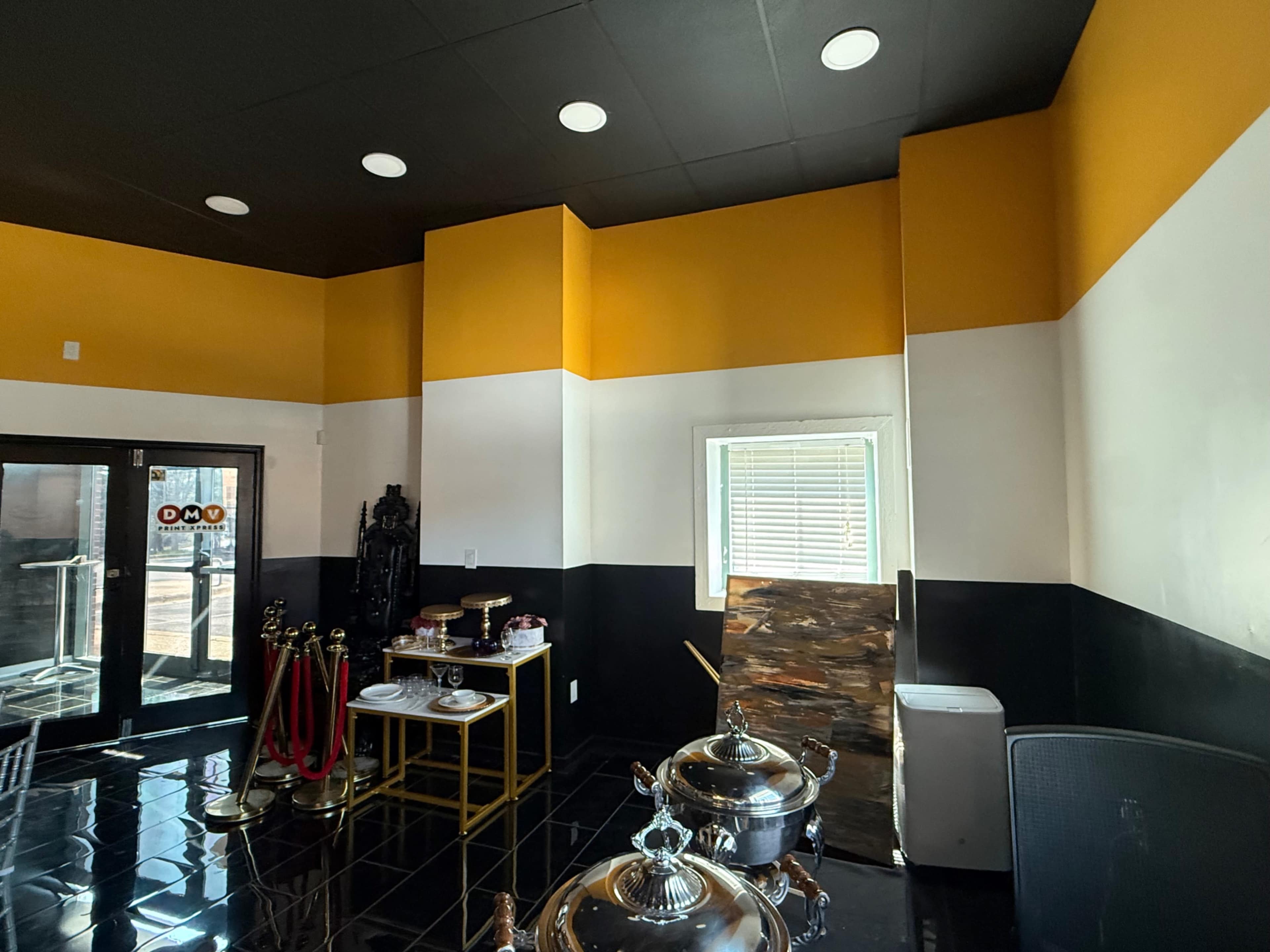 The image shows the interior of a restaurant featuring black, white, and yellow wall colors, a window, and some furniture including a buffet table and a small serving cart.