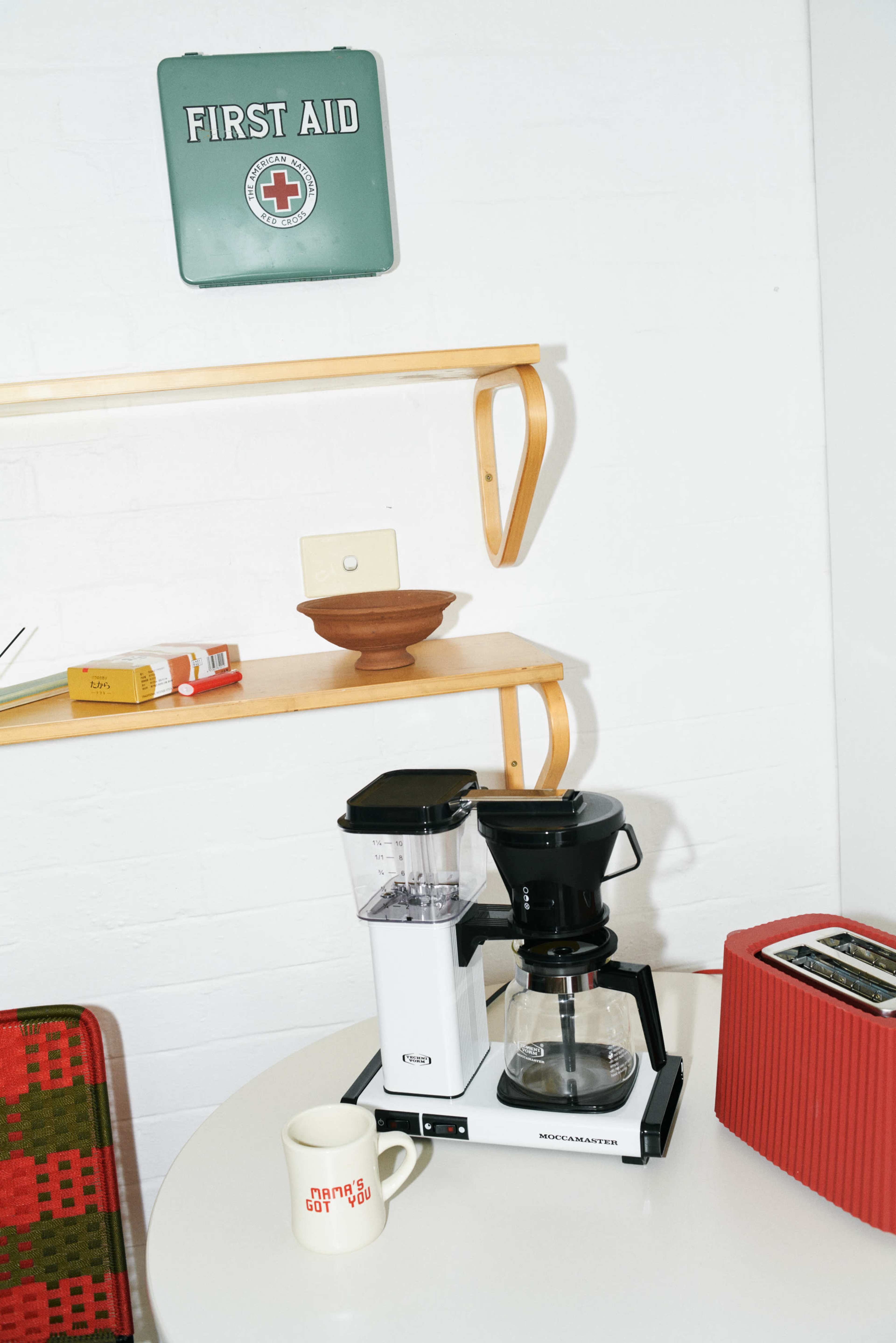 A coffee maker and toaster are positioned on a small round table, with a first aid sign hanging on the wall above a wooden shelf displaying a bowl and other items.