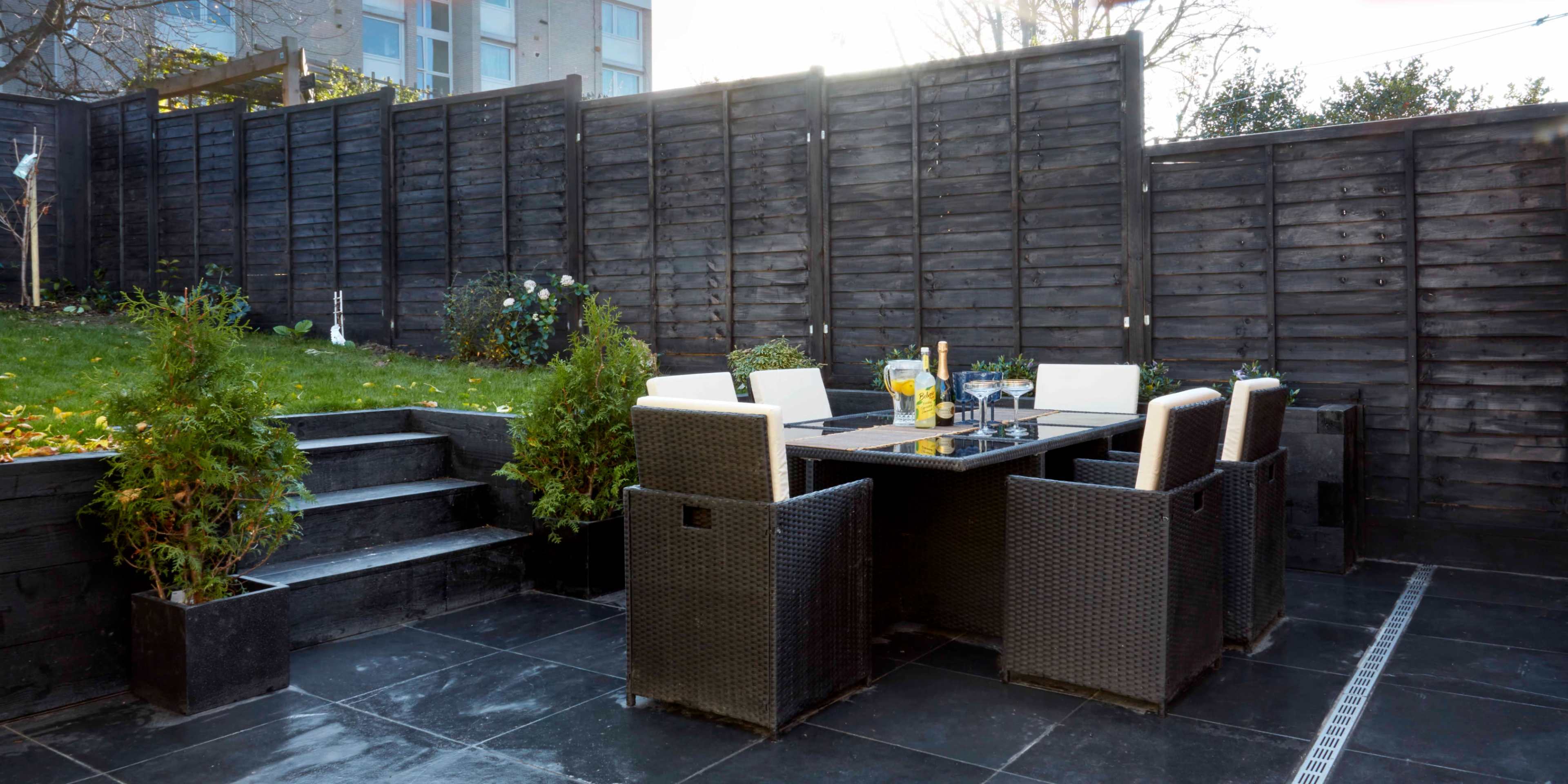 A patio area features a table set with chairs, surrounded by a landscaped yard and a wooden fence.
