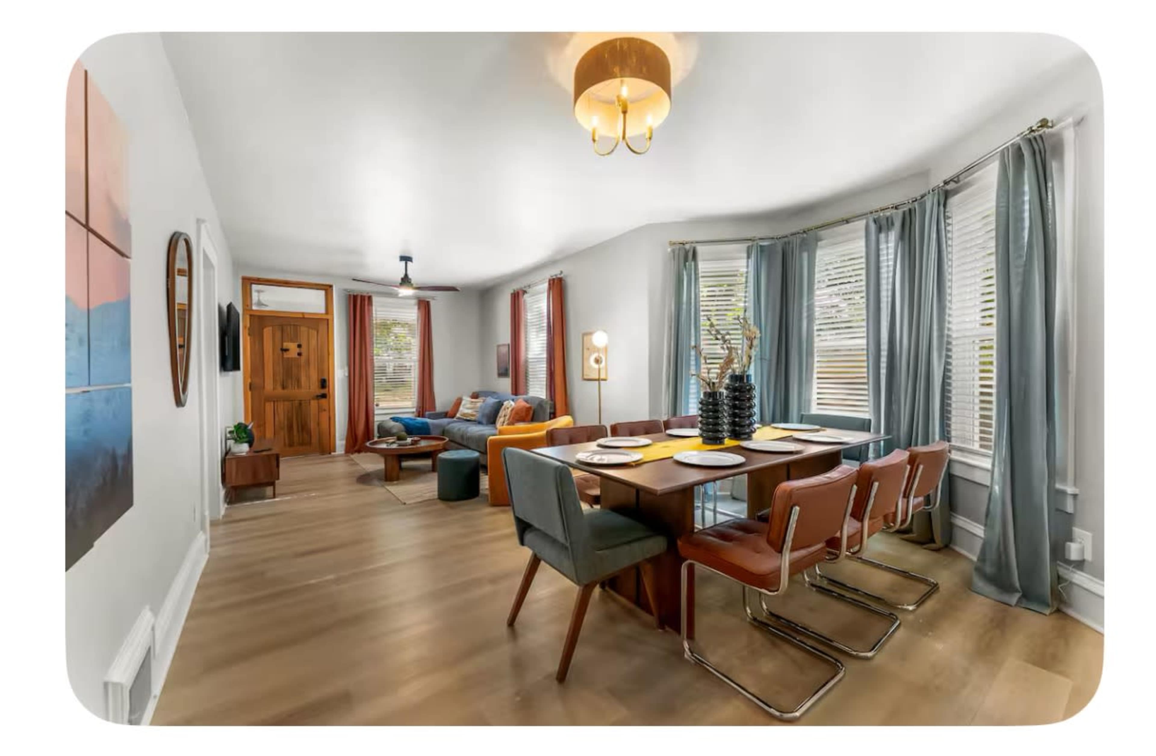 The image shows a modern living space featuring a dining table set for four, a cozy seating area with a blue couch, and large windows decorated with light blue curtains.