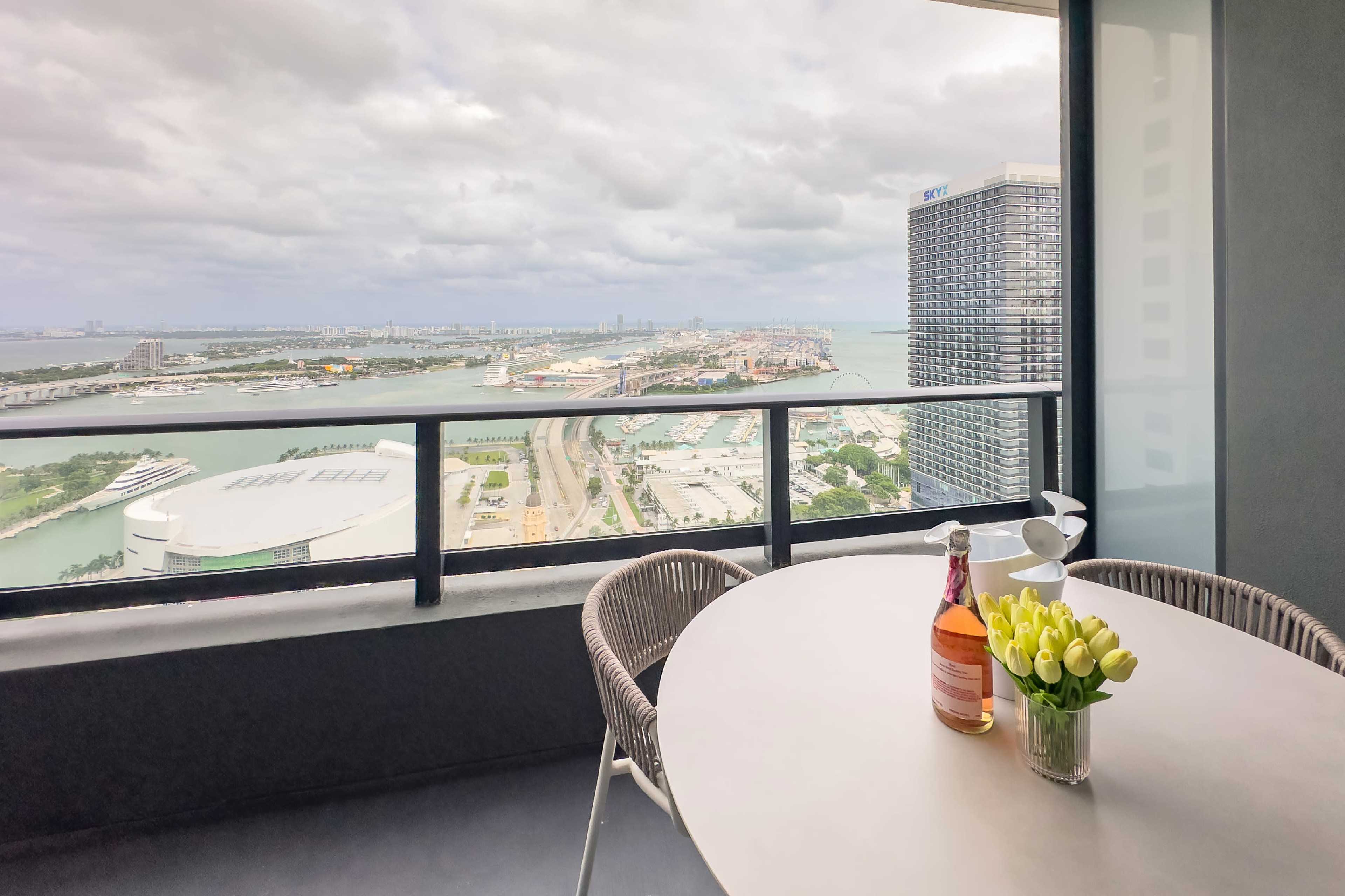 A balcony table set with a bottle of rose wine and a vase of yellow tulips overlooks a cityscape and waterfront.