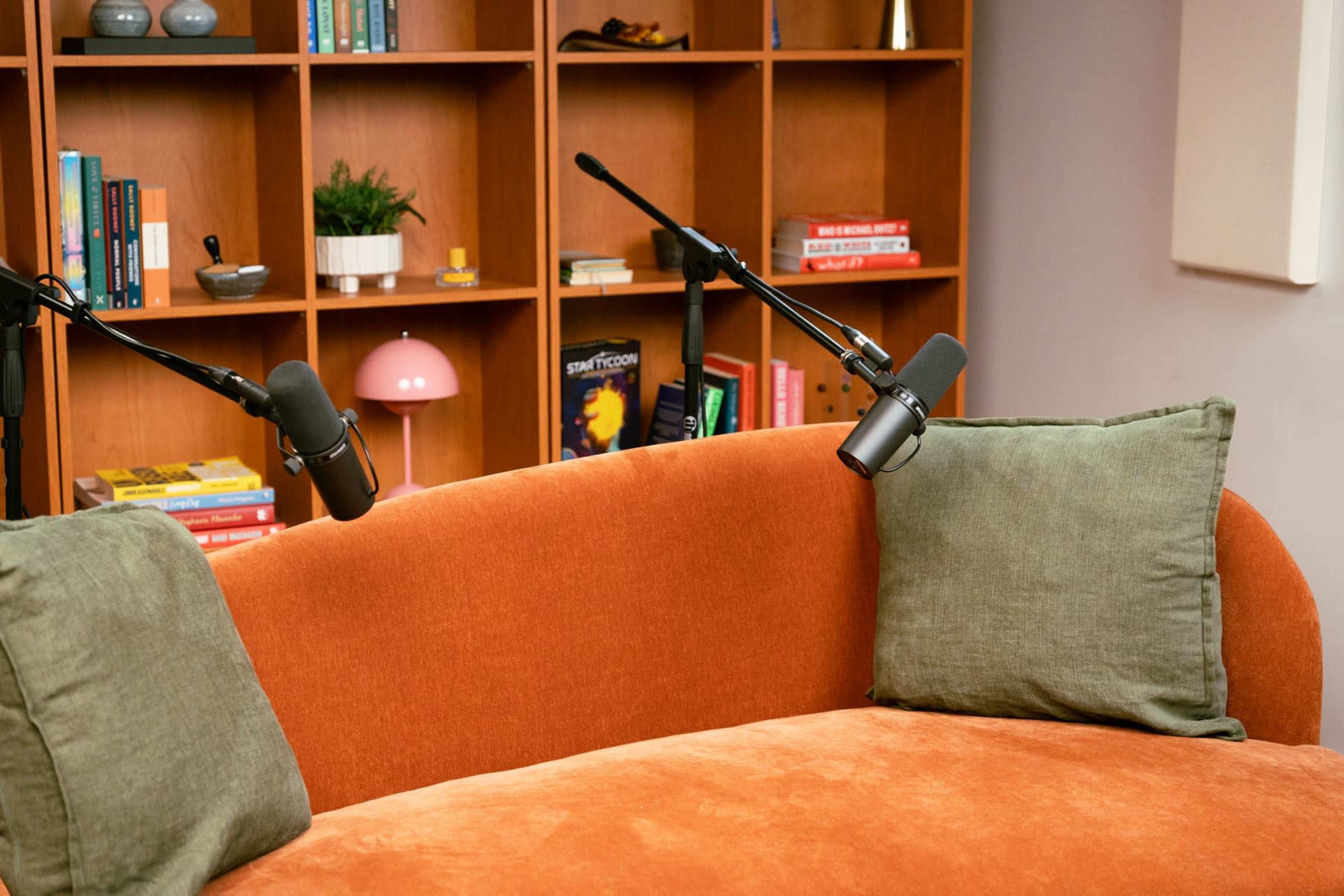 A cozy orange sofa with green cushions is positioned in front of a wooden bookshelf filled with books and decorative items, with two microphones mounted on stands pointing toward the seating area.