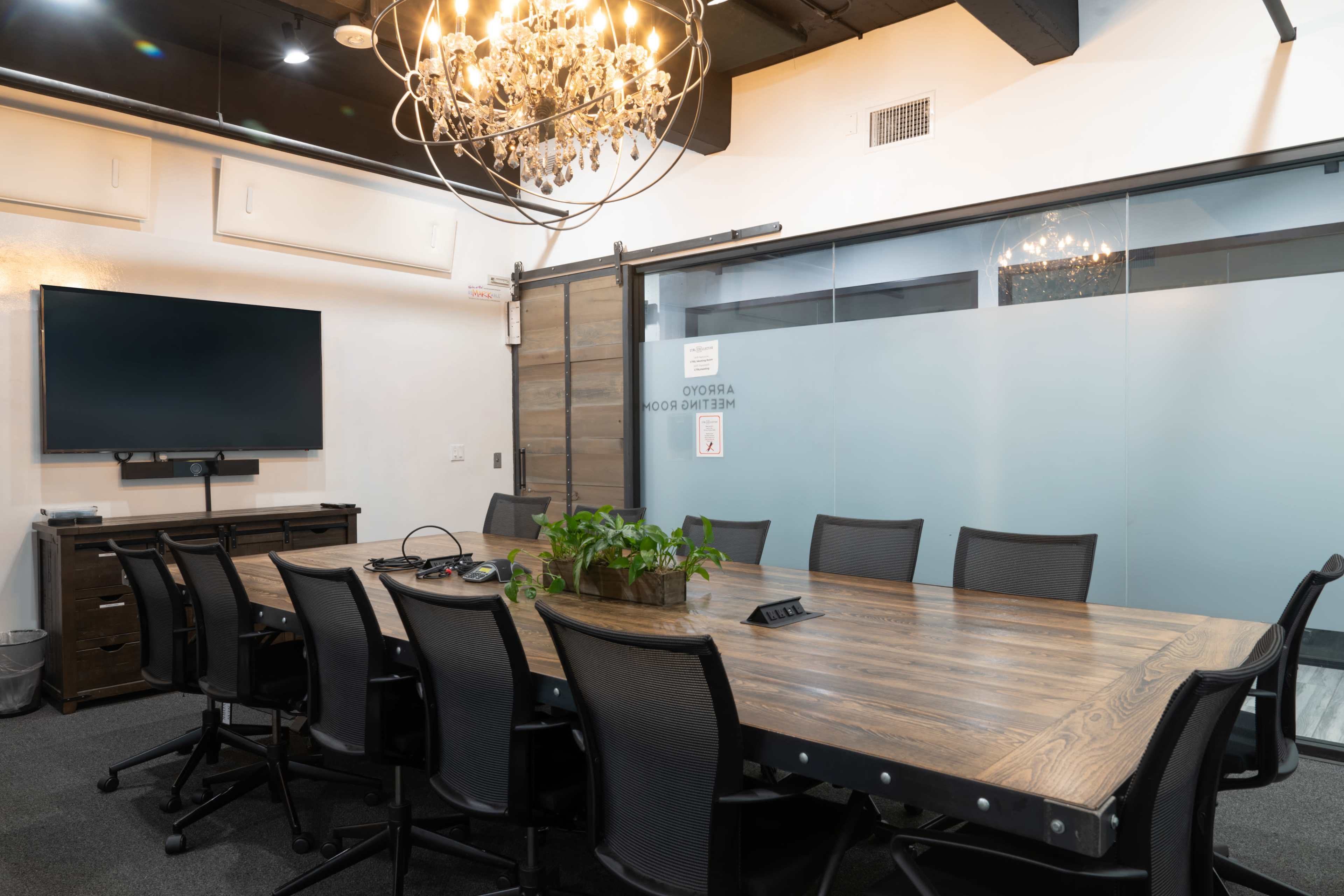 The image shows a conference room with a large wooden table surrounded by black chairs, a wall-mounted television, and a chandelier overhead.