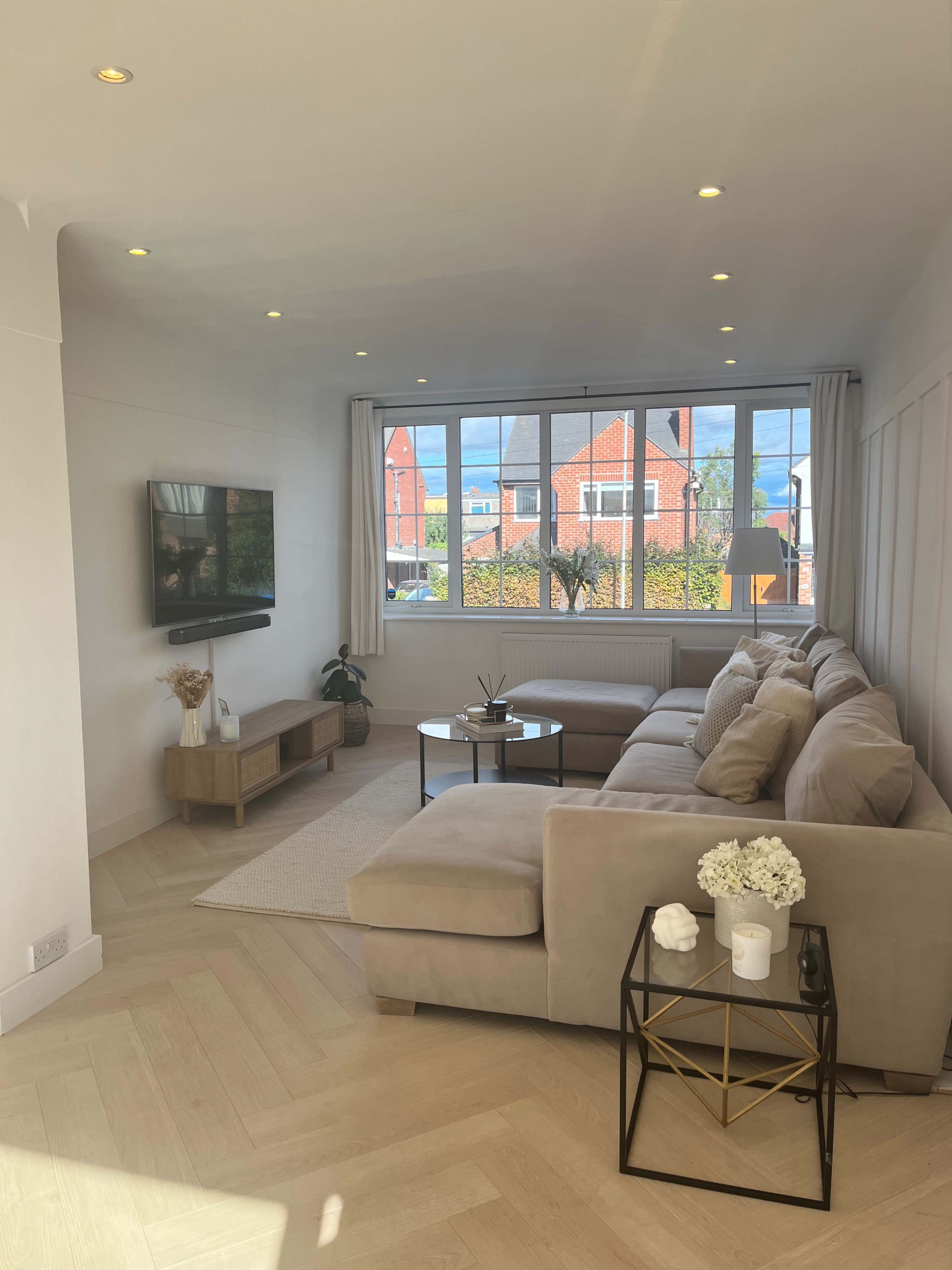 A modern living room with a large sectional sofa, a coffee table, a television mounted on the wall, and large windows allowing natural light to enter.