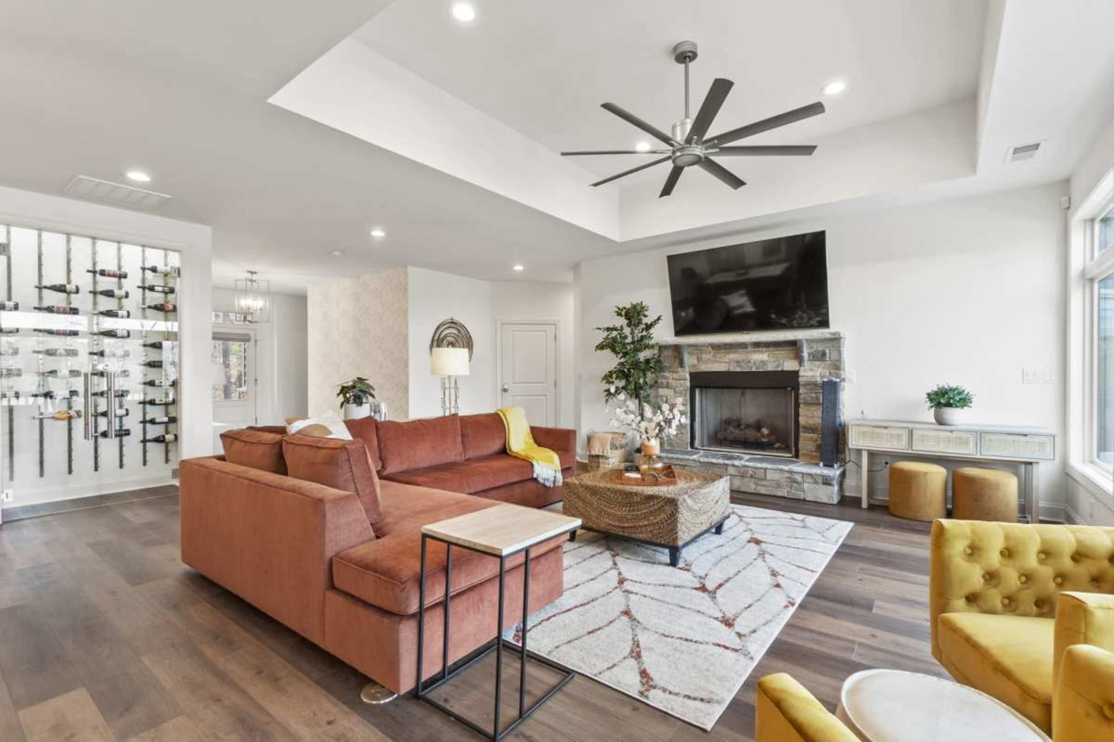 The image shows a spacious living room featuring a large orange sectional sofa, a stone fireplace with a mounted television above it, and a wine wall on the left side.