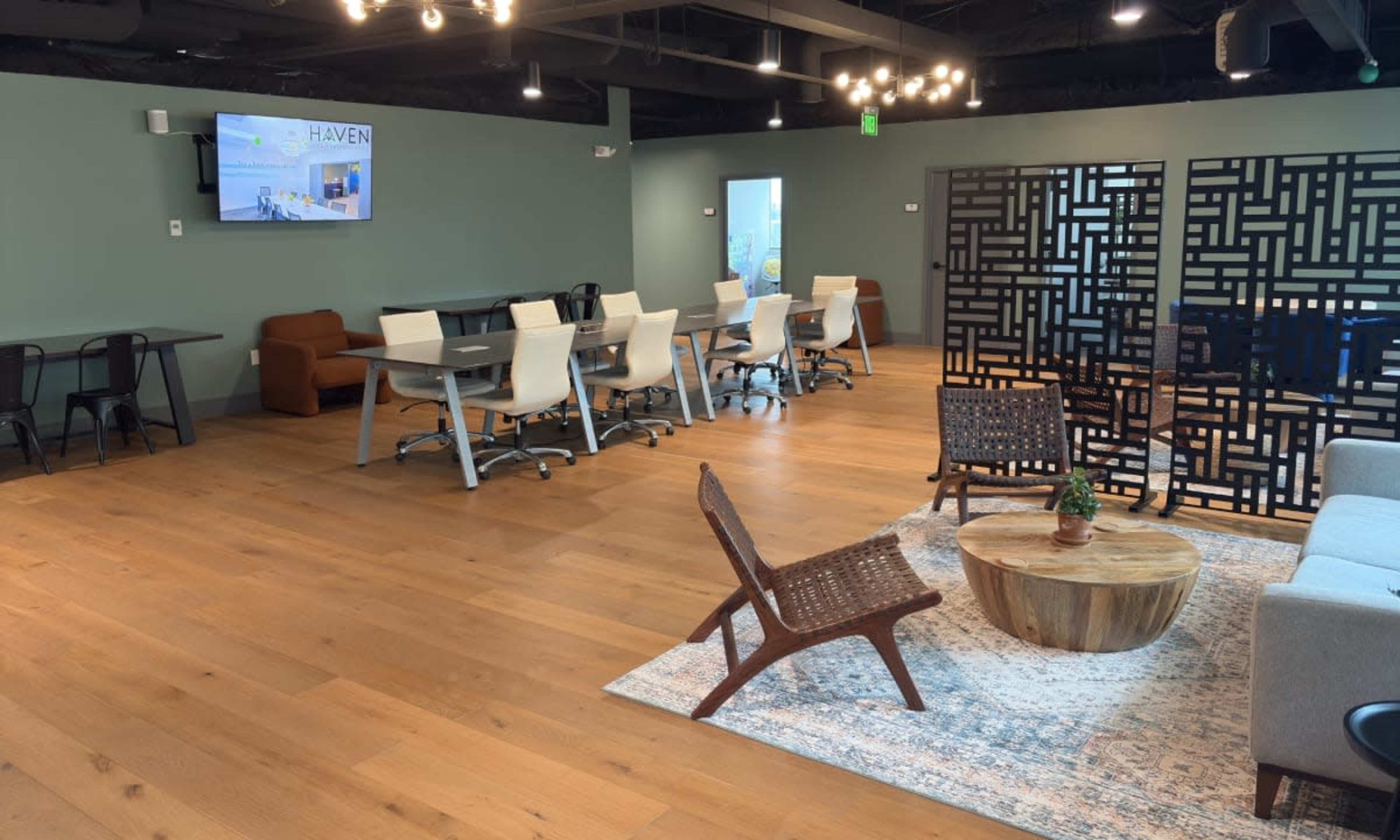 The image shows a modern and spacious communal workspace with a mix of seating options, including tables and chairs, lounge areas, and a television on the wall.