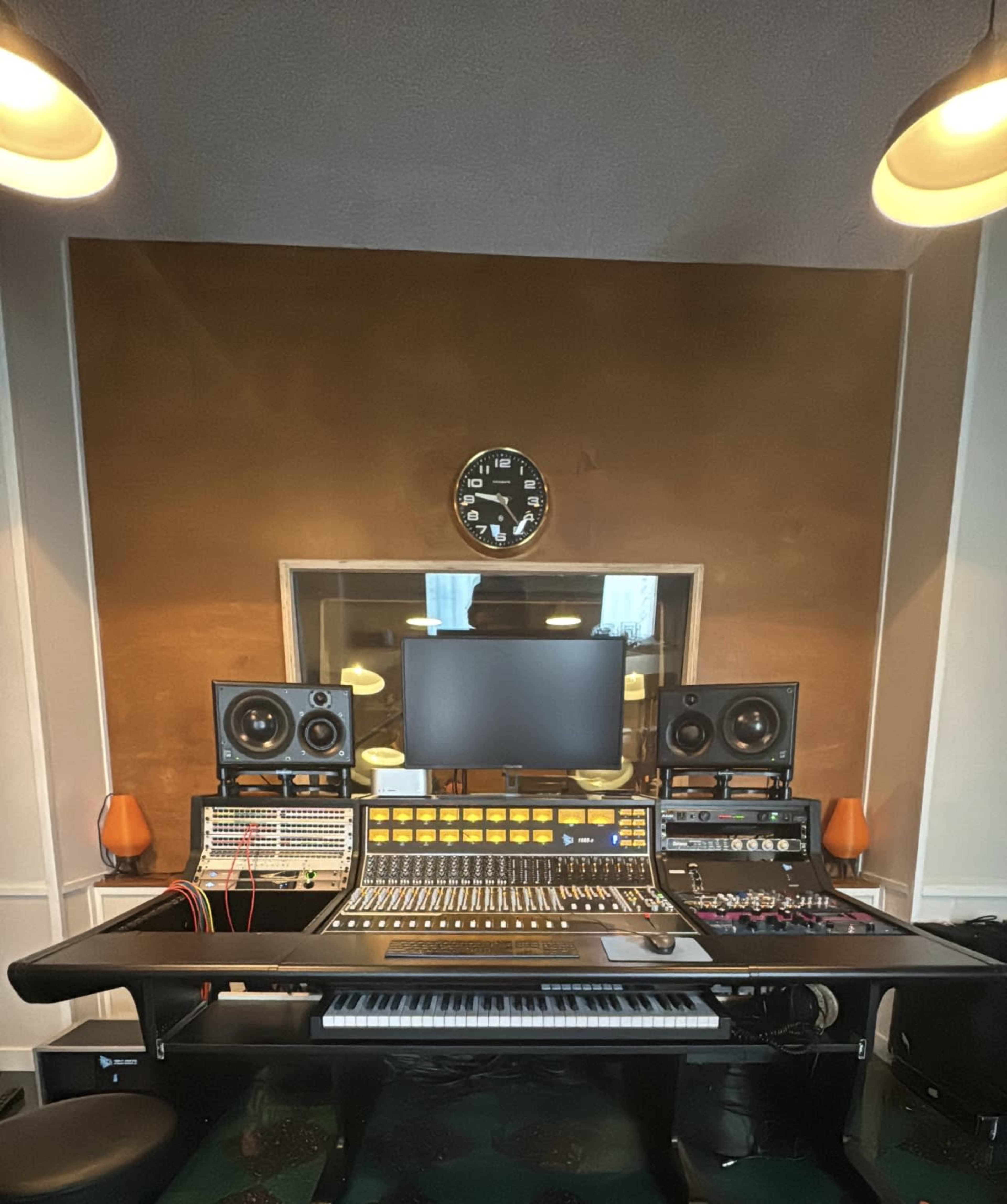 A music studio is shown, featuring a large mixing console, computer monitor, speakers, and various audio equipment arranged in an organized manner.