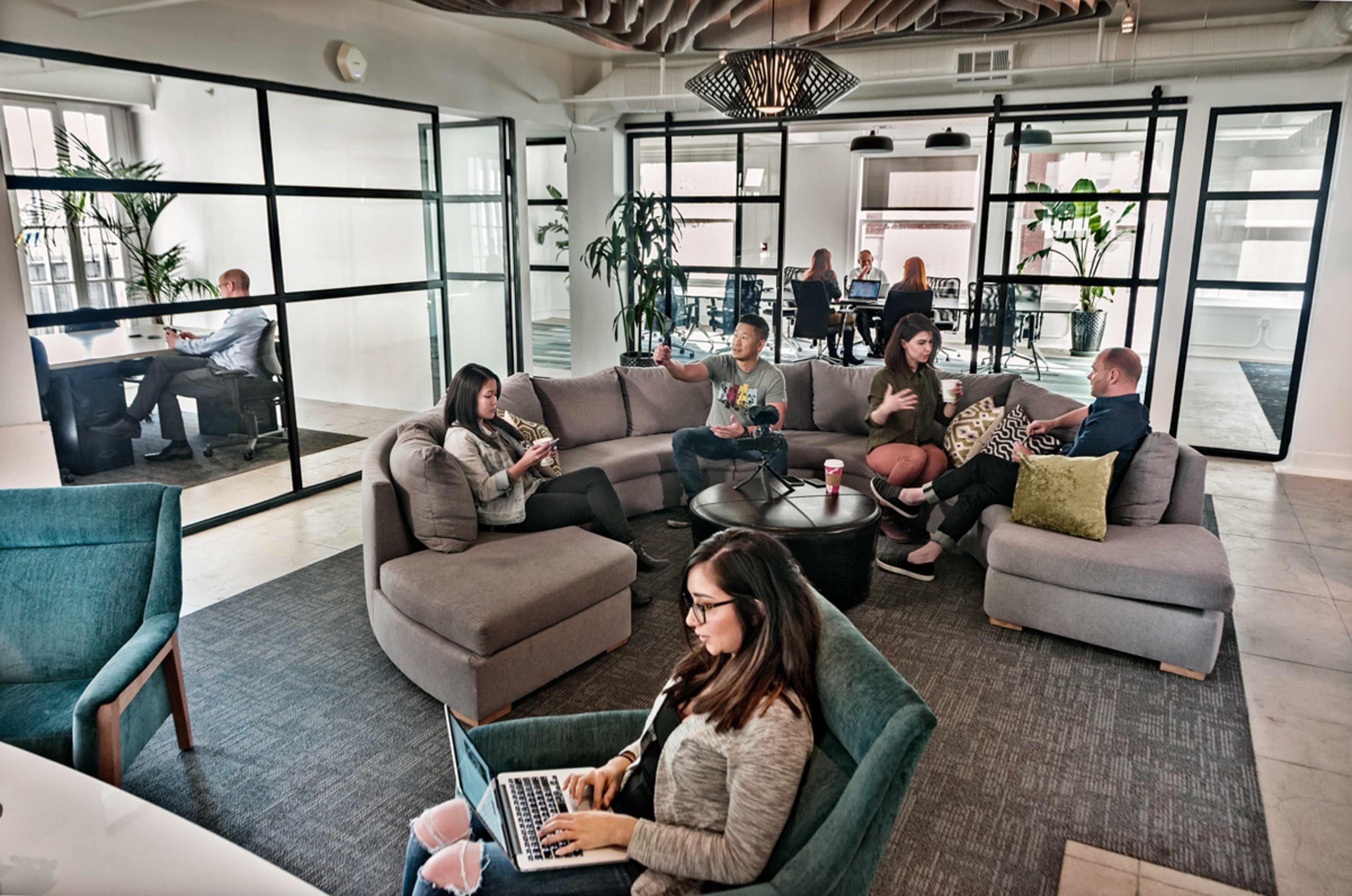 A group of people are working and engaging in conversation in a modern office space with a lounge area and glass meeting rooms.