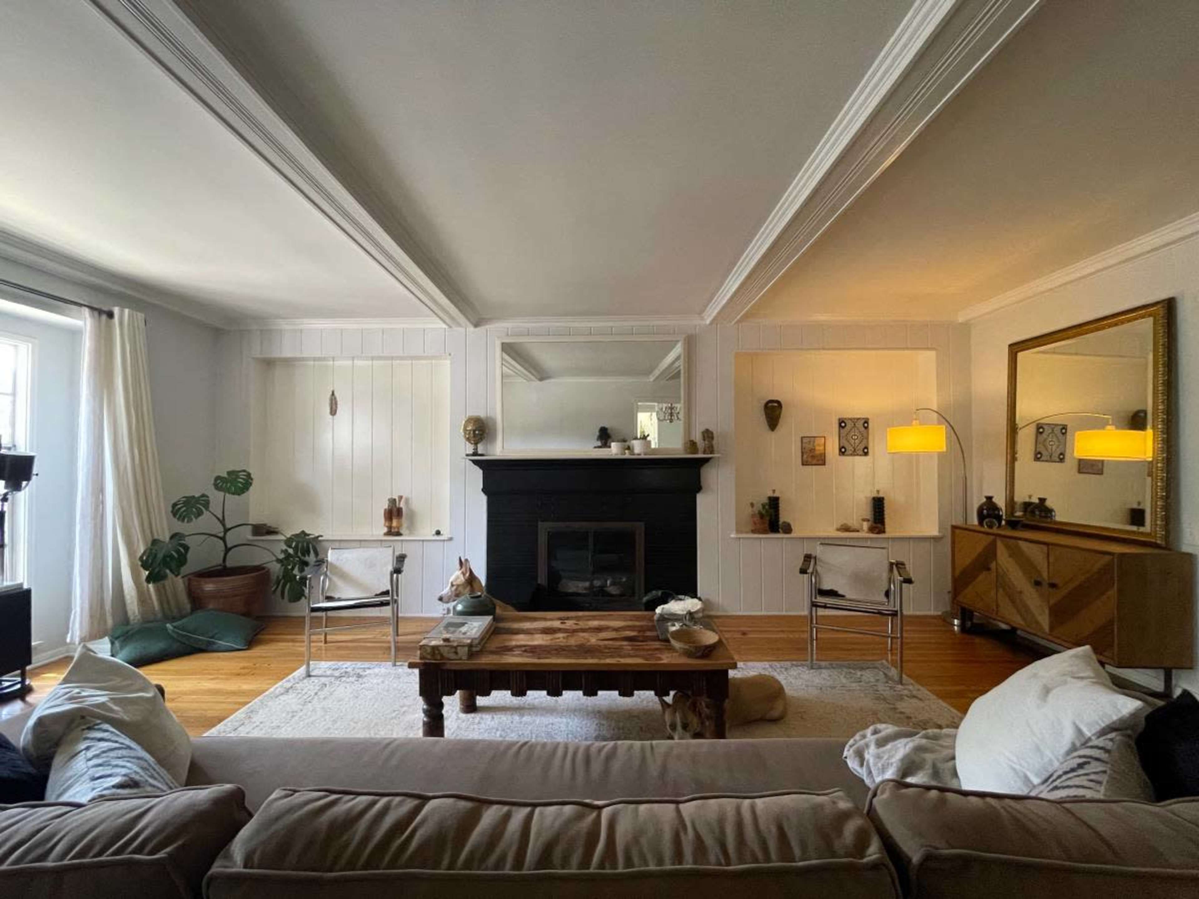 The living room features a central coffee table surrounded by seating, a fireplace with decorative elements, and large windows allowing natural light.