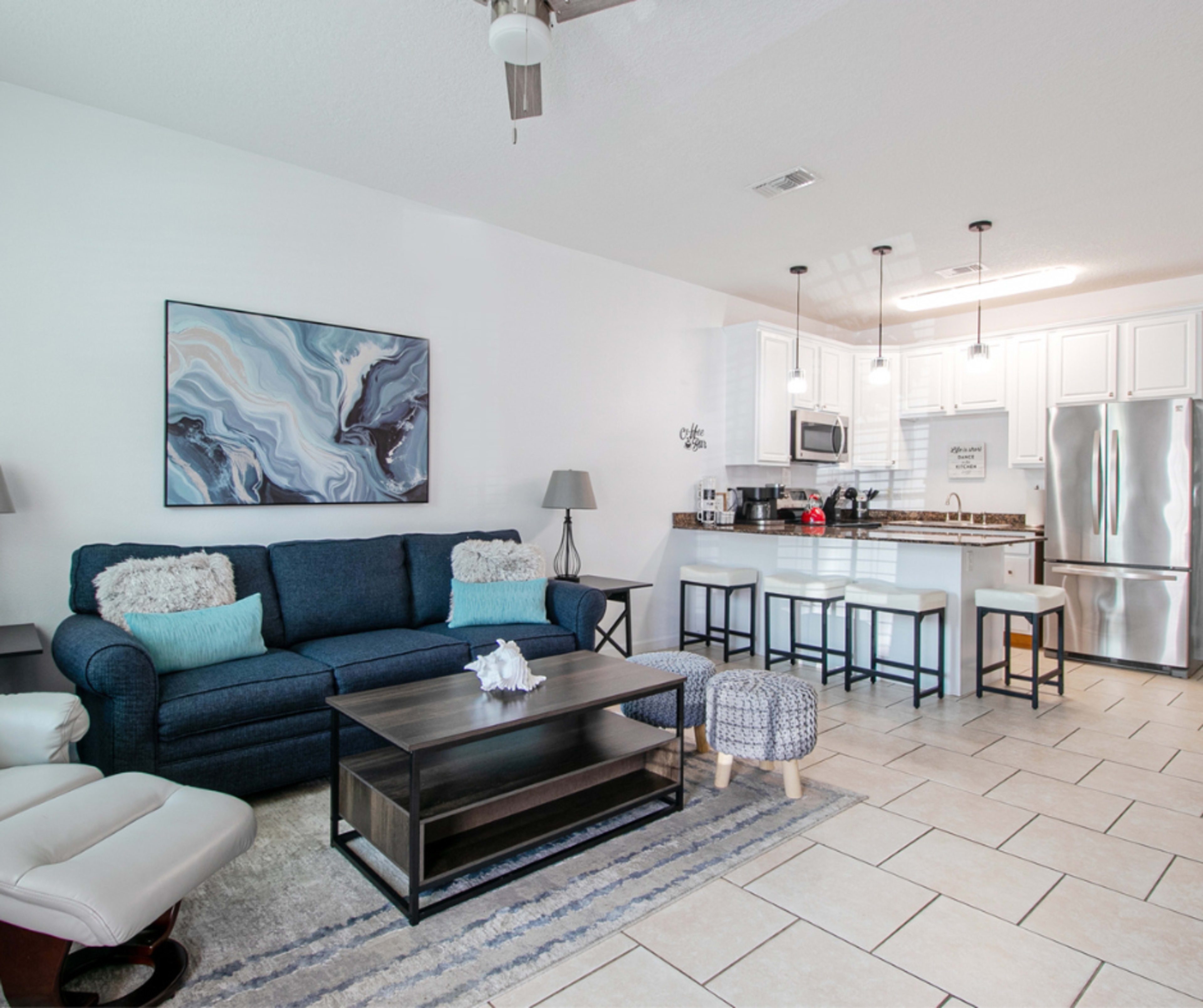 The image shows a modern living area with a blue sofa, a coffee table, and an open kitchen featuring white cabinets and stainless steel appliances.