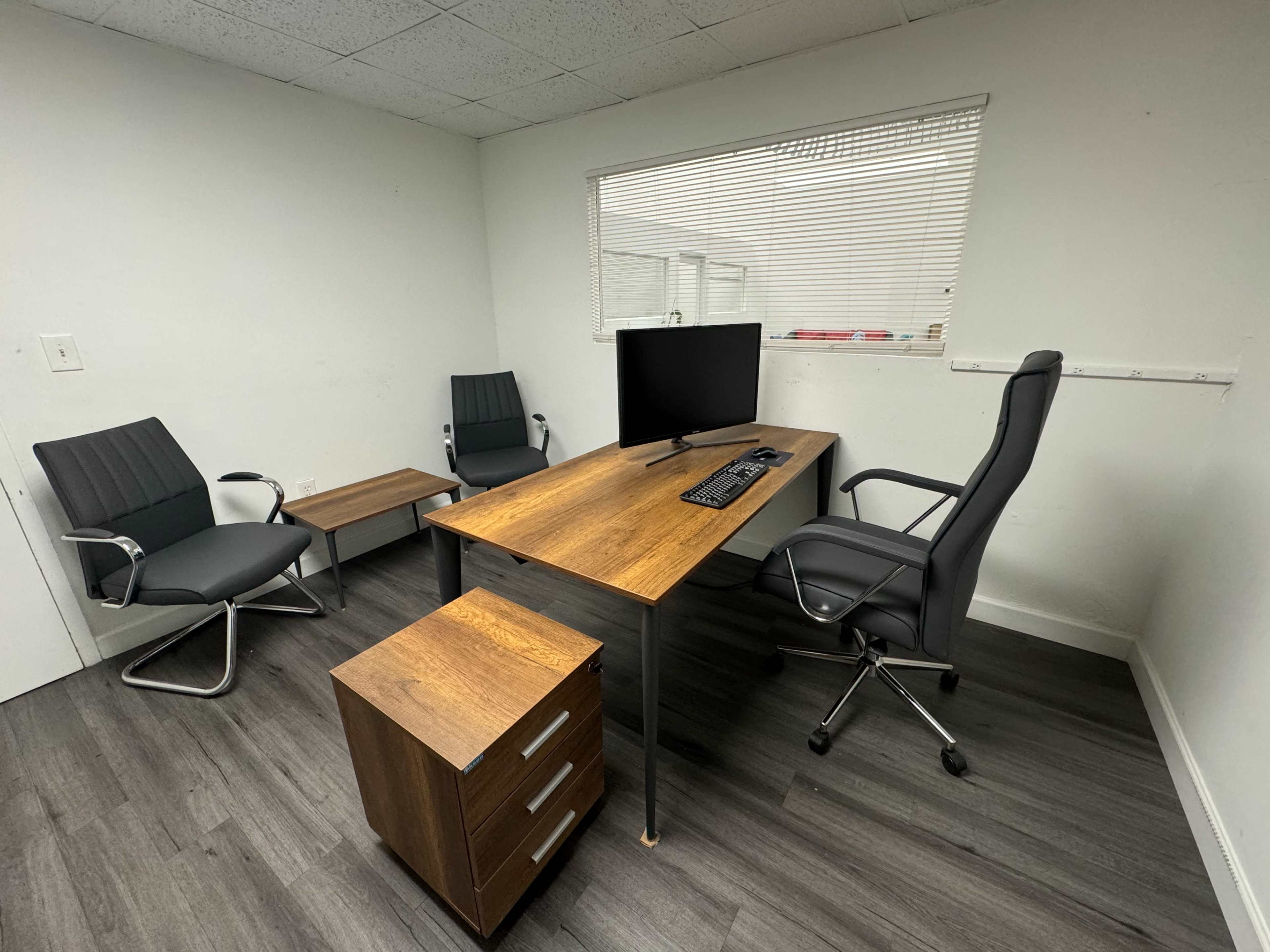 A minimalist office space with a wooden desk, a computer monitor, three chairs, a side table, and a storage unit.