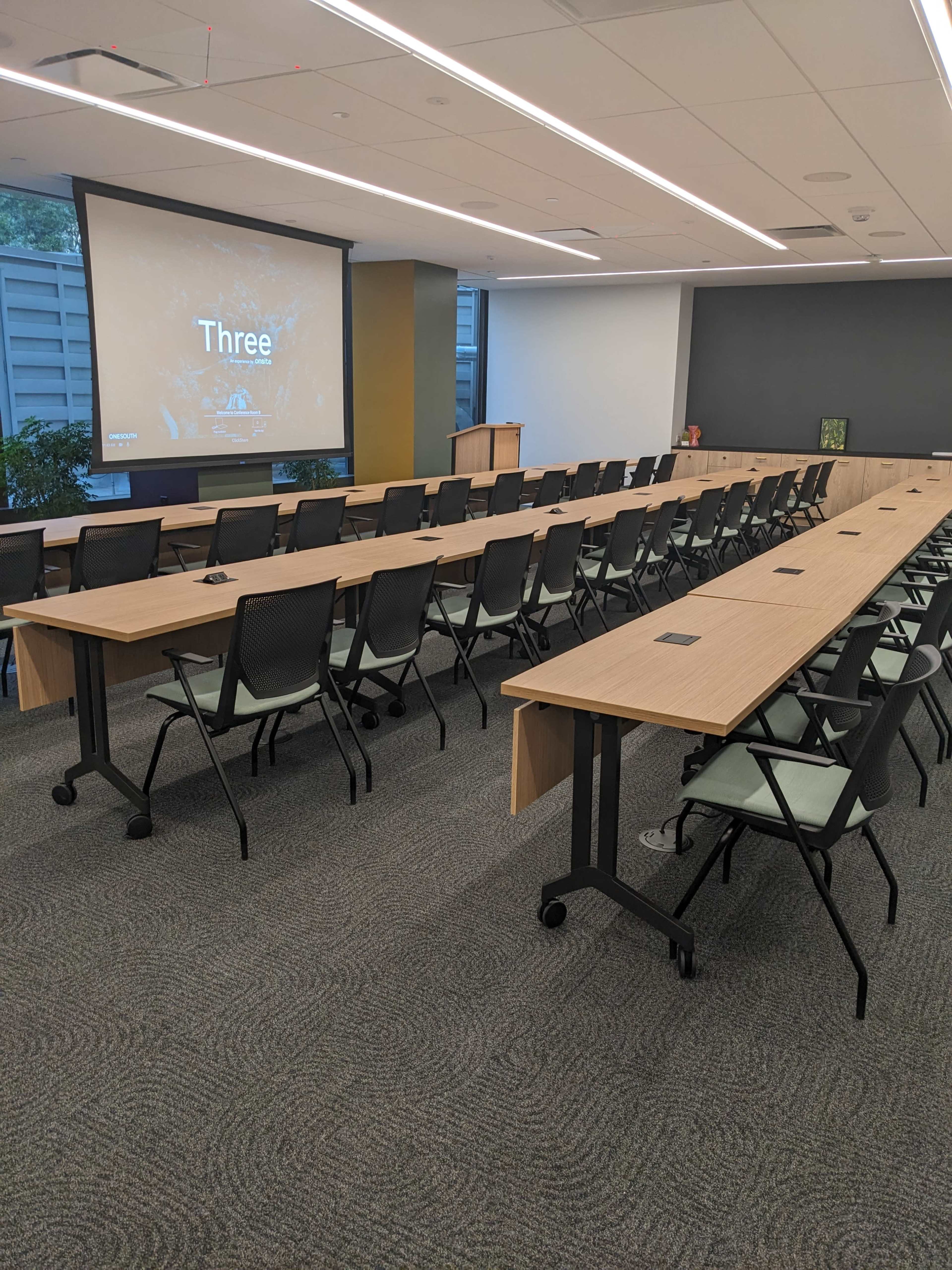 The image shows a modern conference room with several rows of tables and chairs arranged for a meeting, featuring a presentation screen at the front.