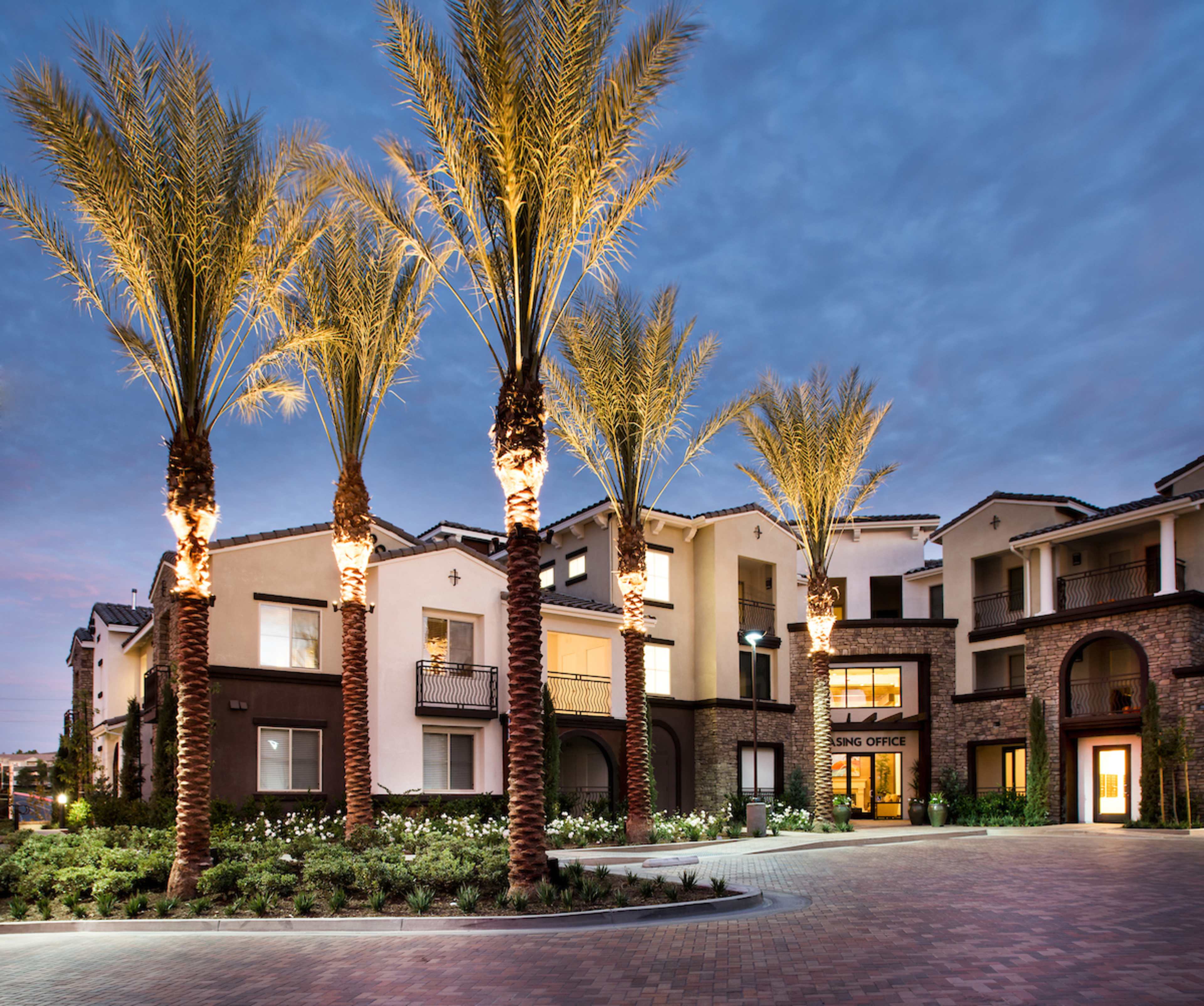 A modern apartment complex with palm trees illuminated at night, surrounded by landscaped gardens.