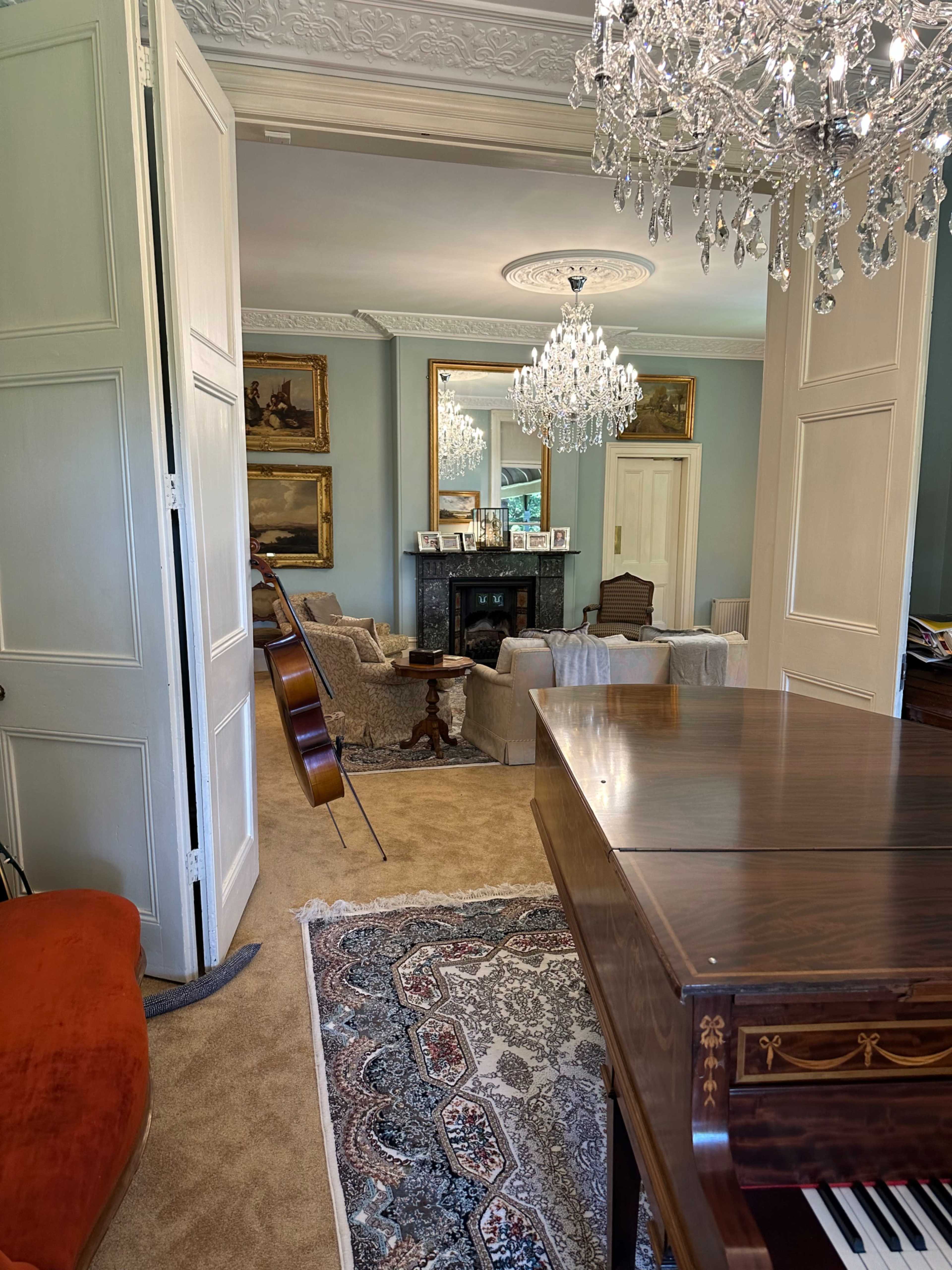 An elegant room with a grand piano, a cello leaning against the wall, and a chandelier above a seating area near a fireplace.