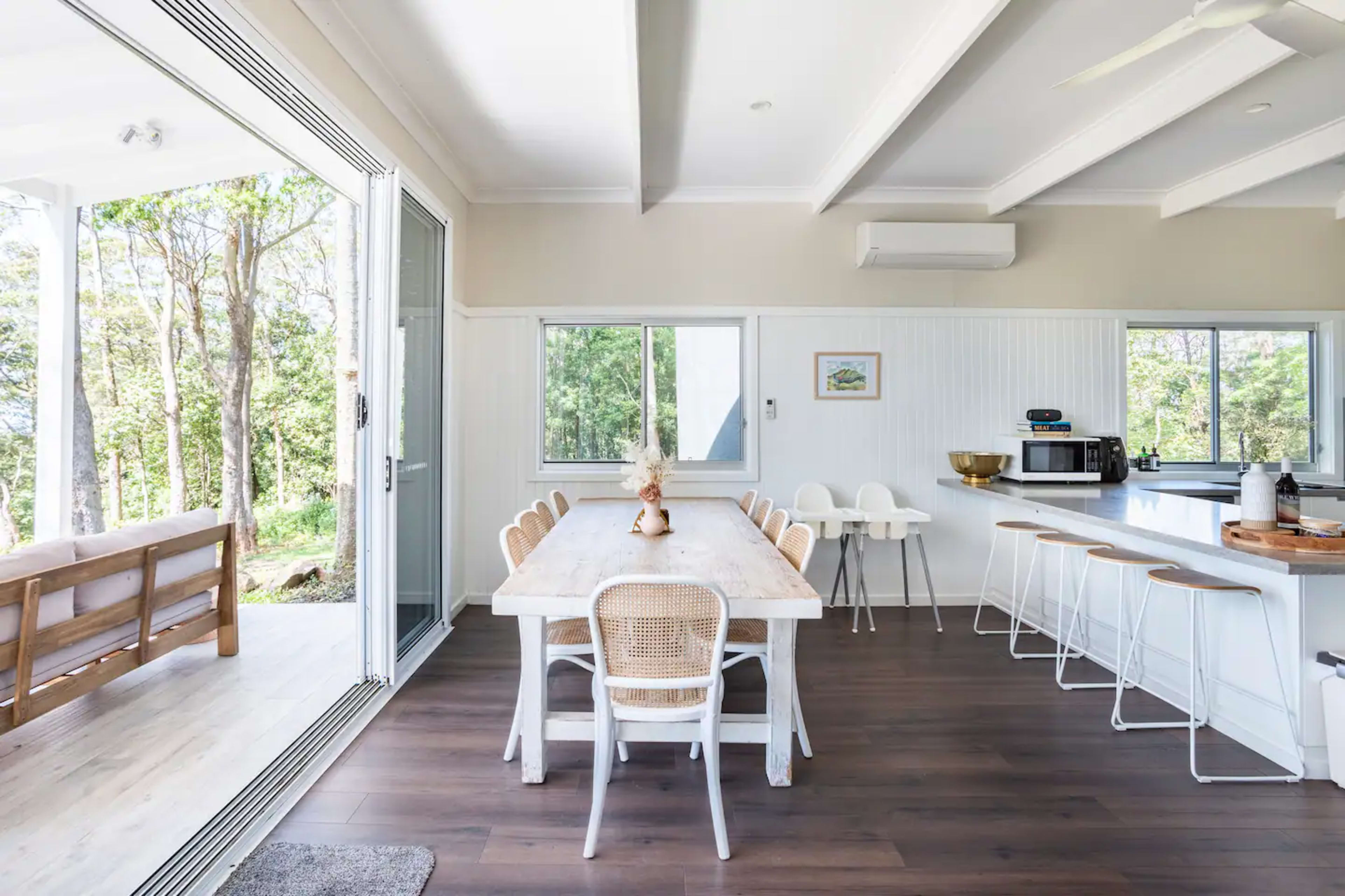 A bright and airy kitchen and dining area features a long wooden table surrounded by chairs, with large windows offering a view of the outdoors.