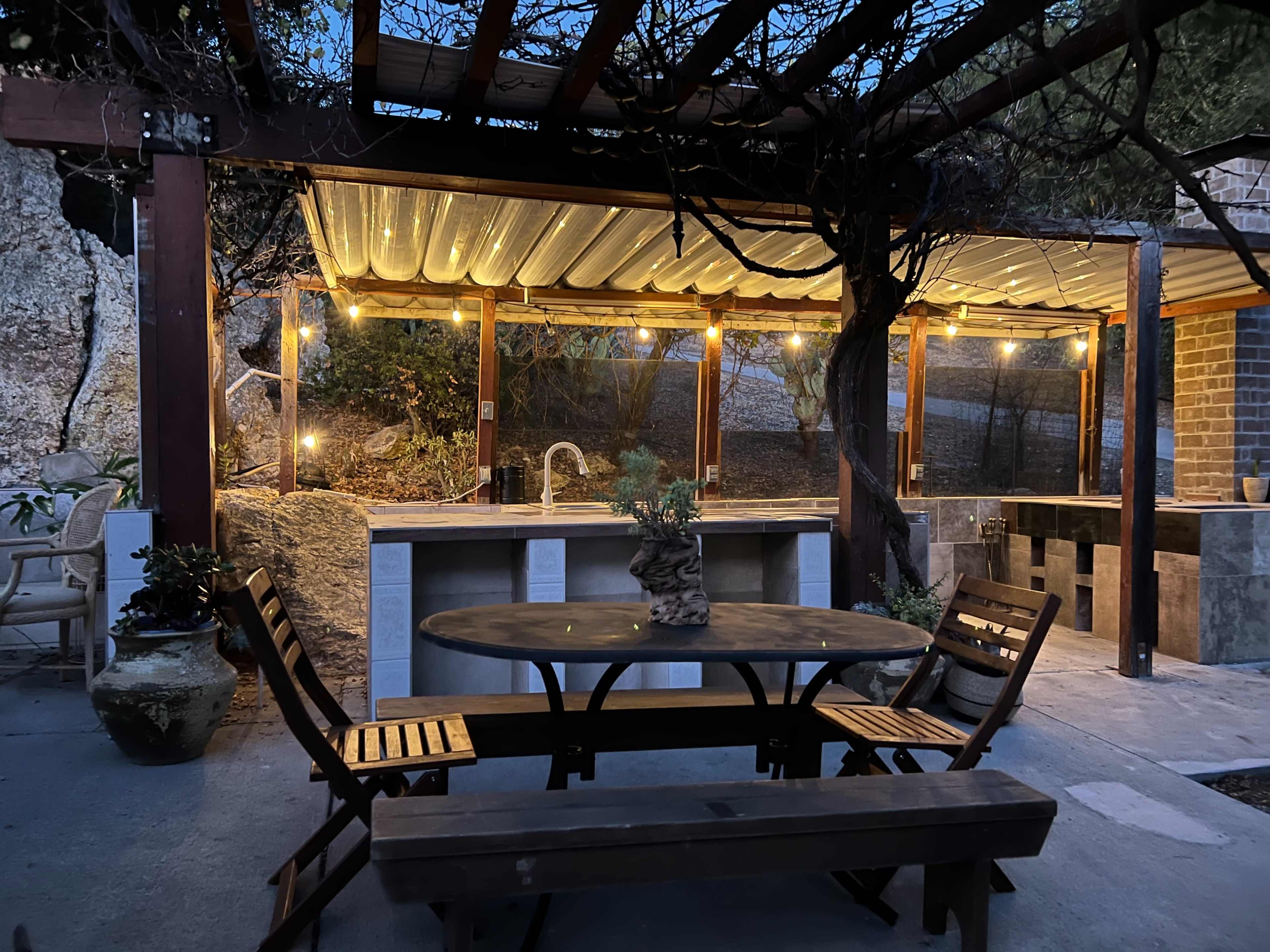 An outdoor kitchen area with a wooden table and benches, covered by a pergola adorned with string lights.