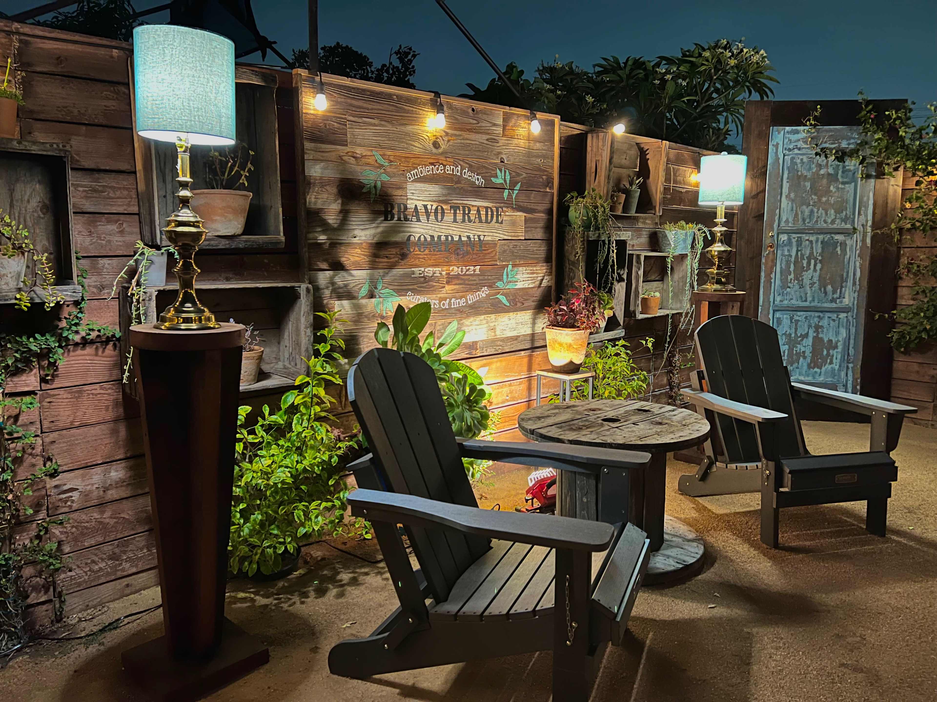 Two wooden Adirondack chairs sit beside a small table under illuminated lamps in a garden setting with a rustic wooden wall and greenery.