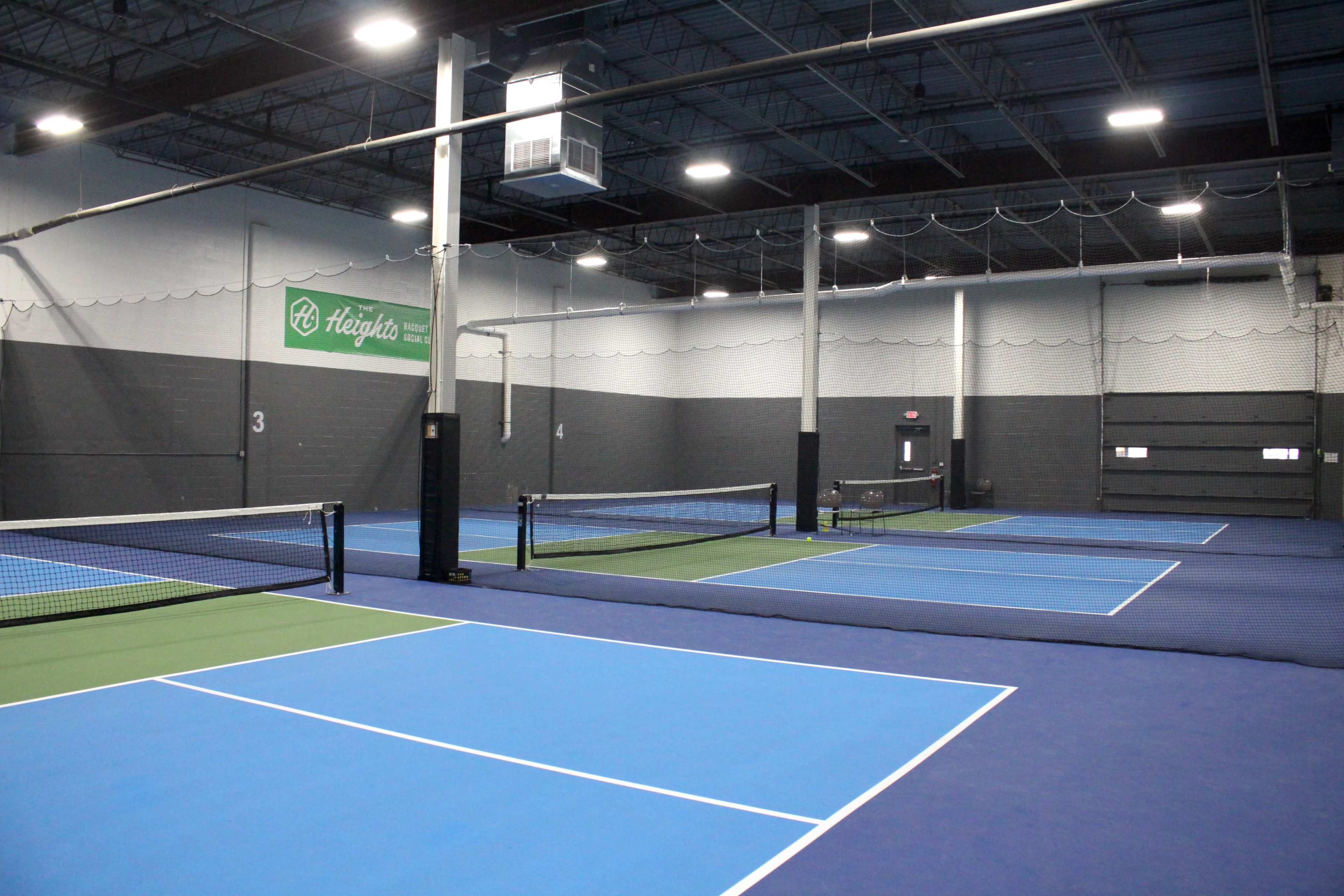 The image shows an indoor tennis facility featuring multiple blue and green courts with nets, and walls painted in dark gray.