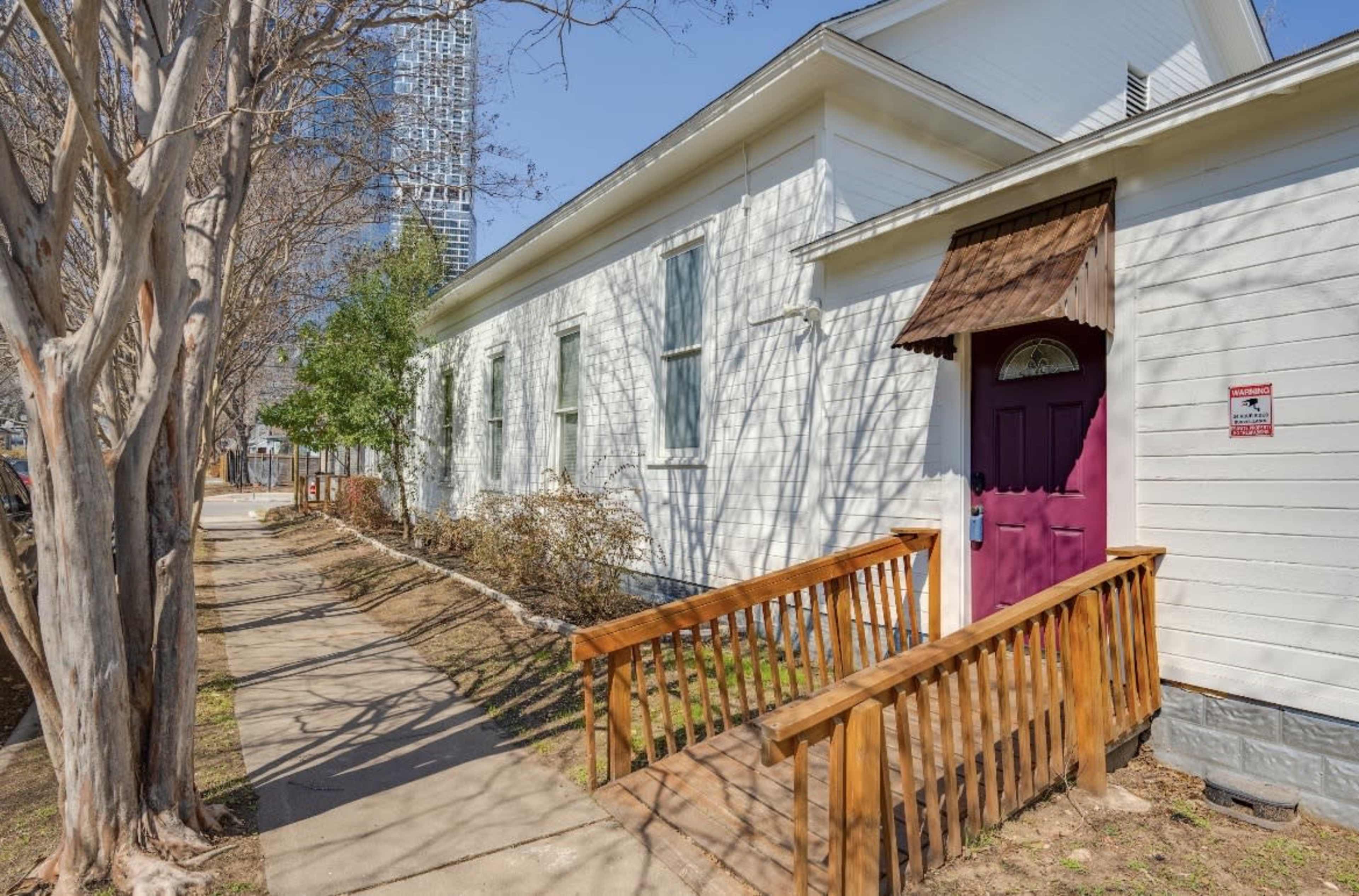 The image shows a white house with a purple door and a wooden ramp, set beside a sidewalk and surrounded by bare trees.