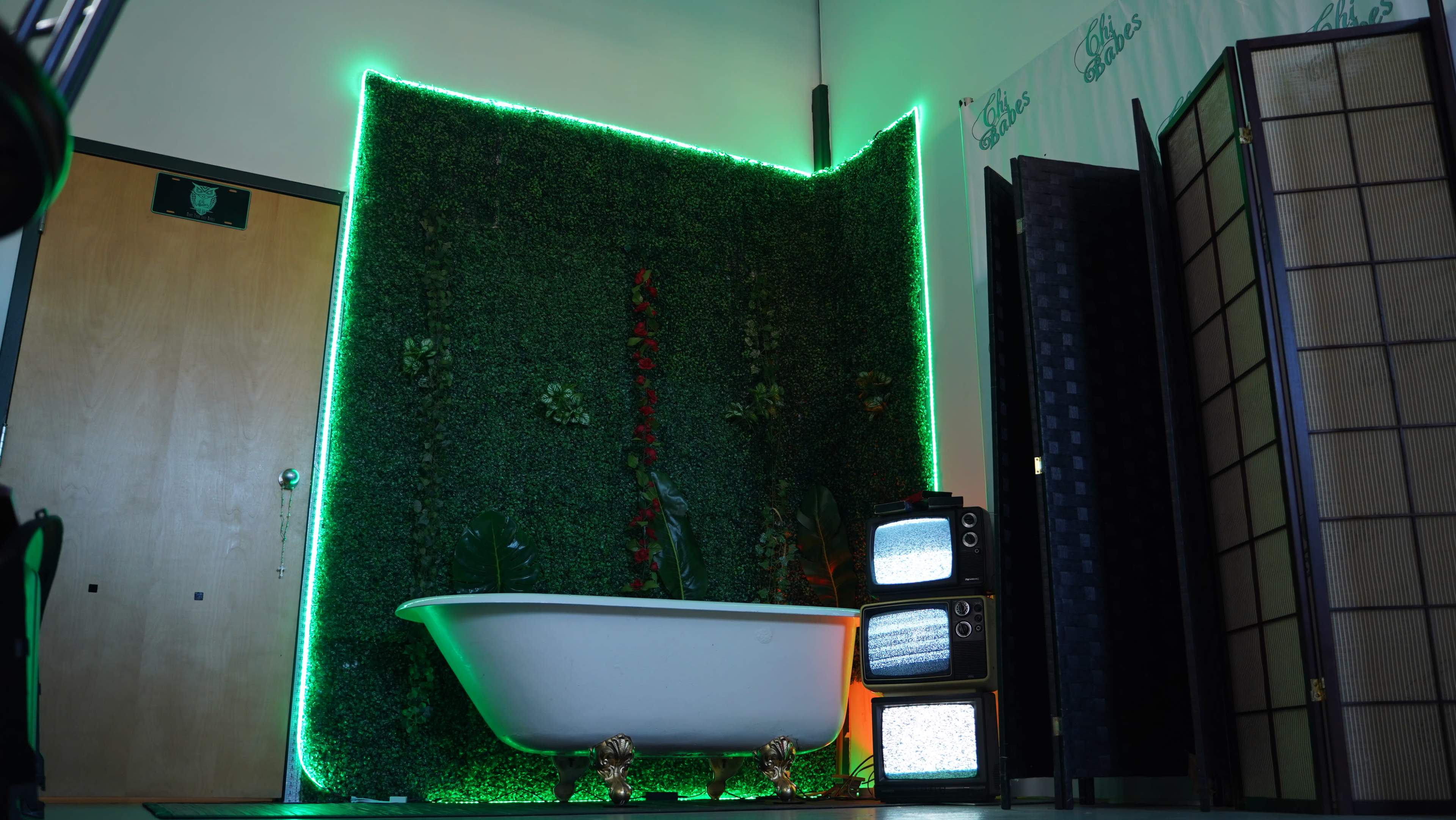 A vintage clawfoot bathtub sits in front of a green wall adorned with plants and illuminated by neon lights, next to a stack of retro television sets.
