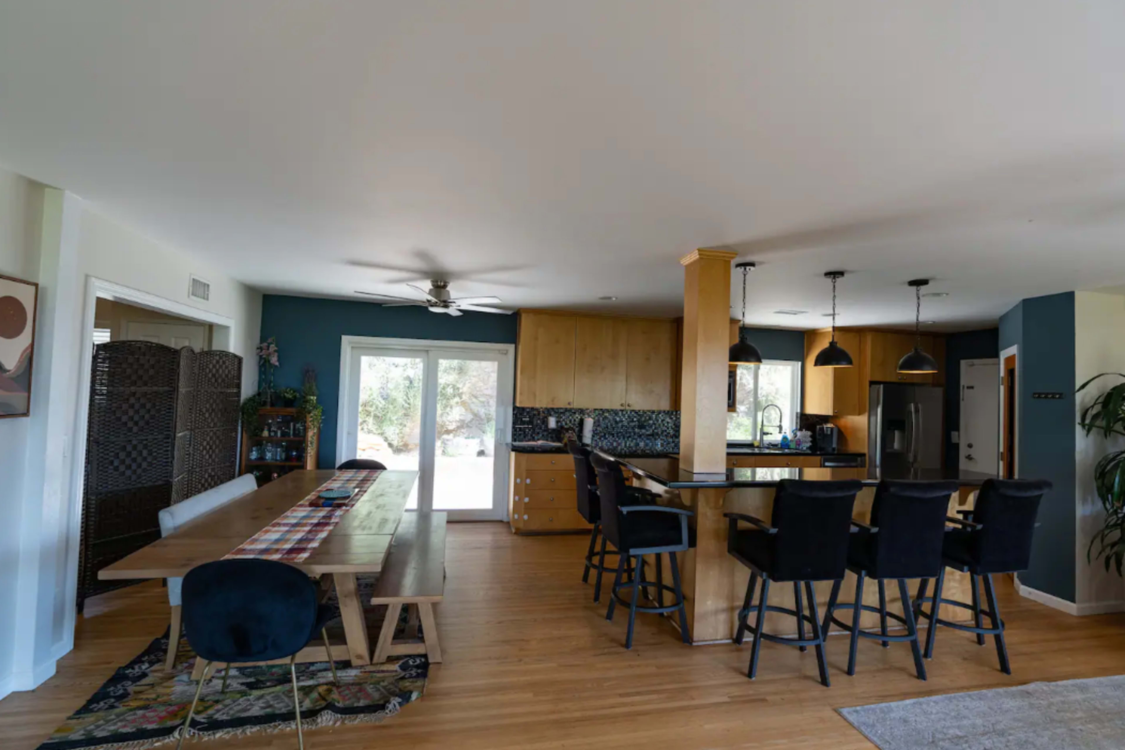 A modern kitchen and dining area with a wooden table, black bar stools, and a large window allowing natural light.