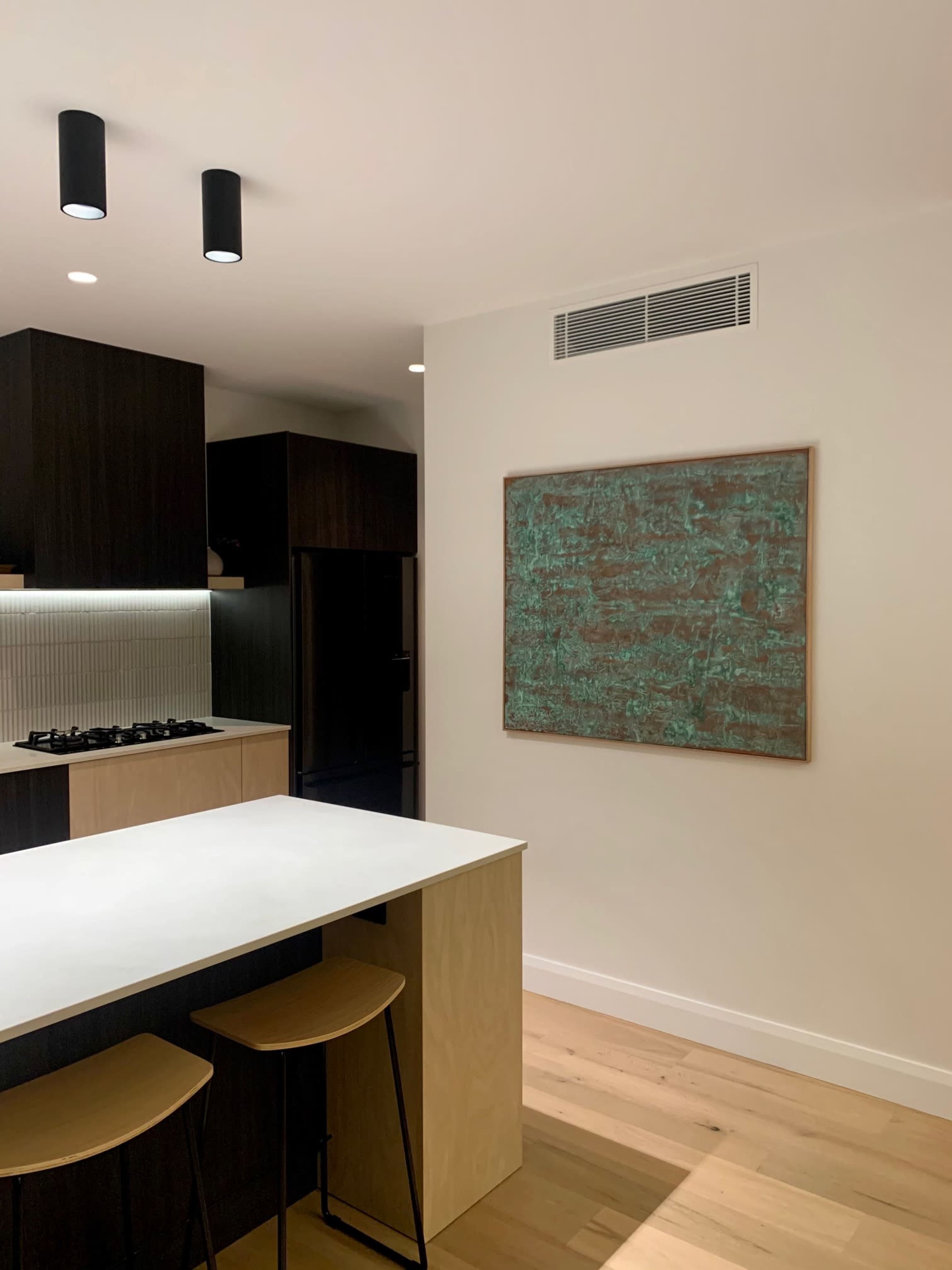 A modern kitchen features dark cabinetry, a white countertop, and a large textured wall art piece.