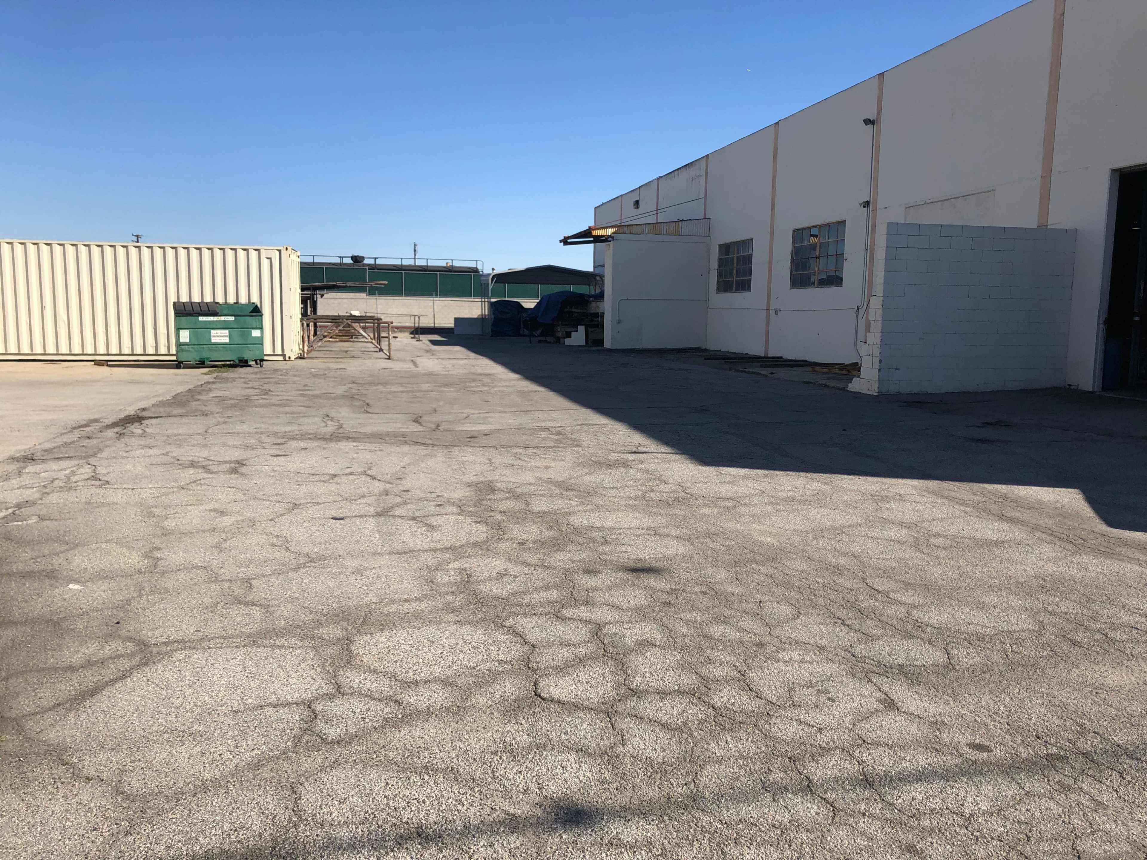 The image shows a large, empty, cracked pavement area next to a warehouse, with a green dumpster and shipping container in the background.