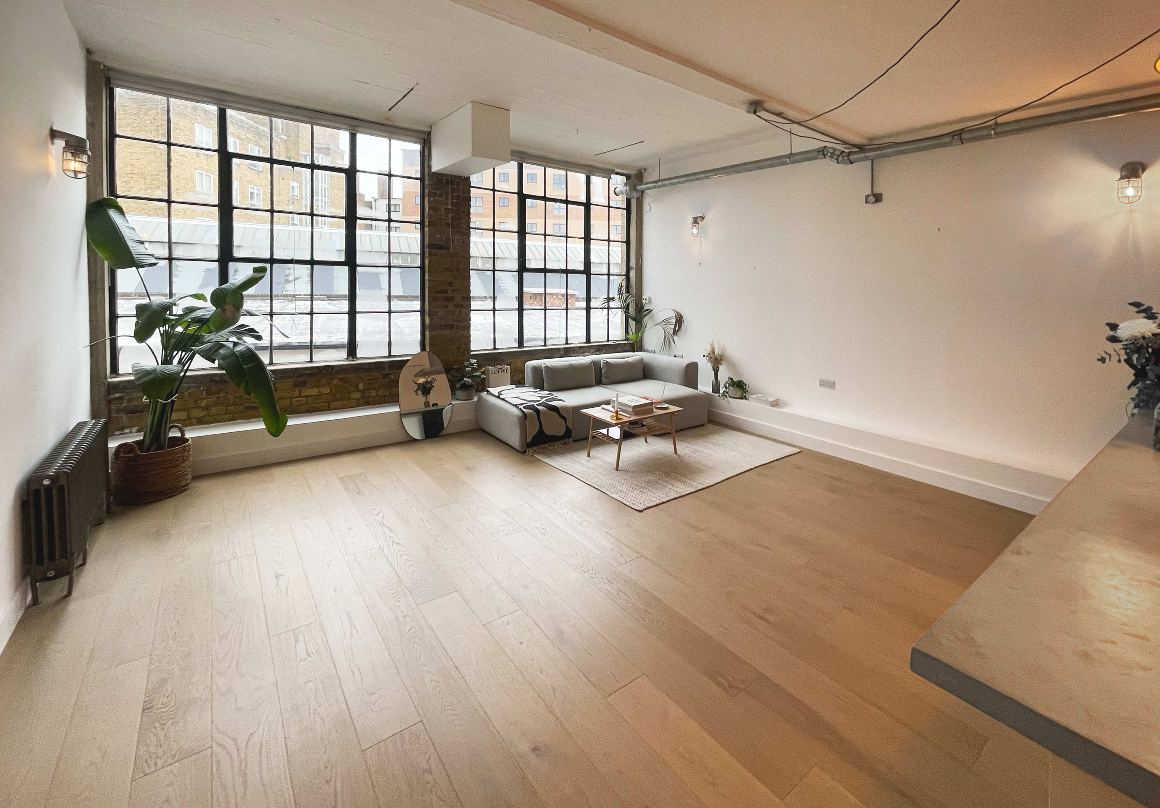 The image shows a minimalist interior living space with large windows, a cozy sofa, and a wooden floor.