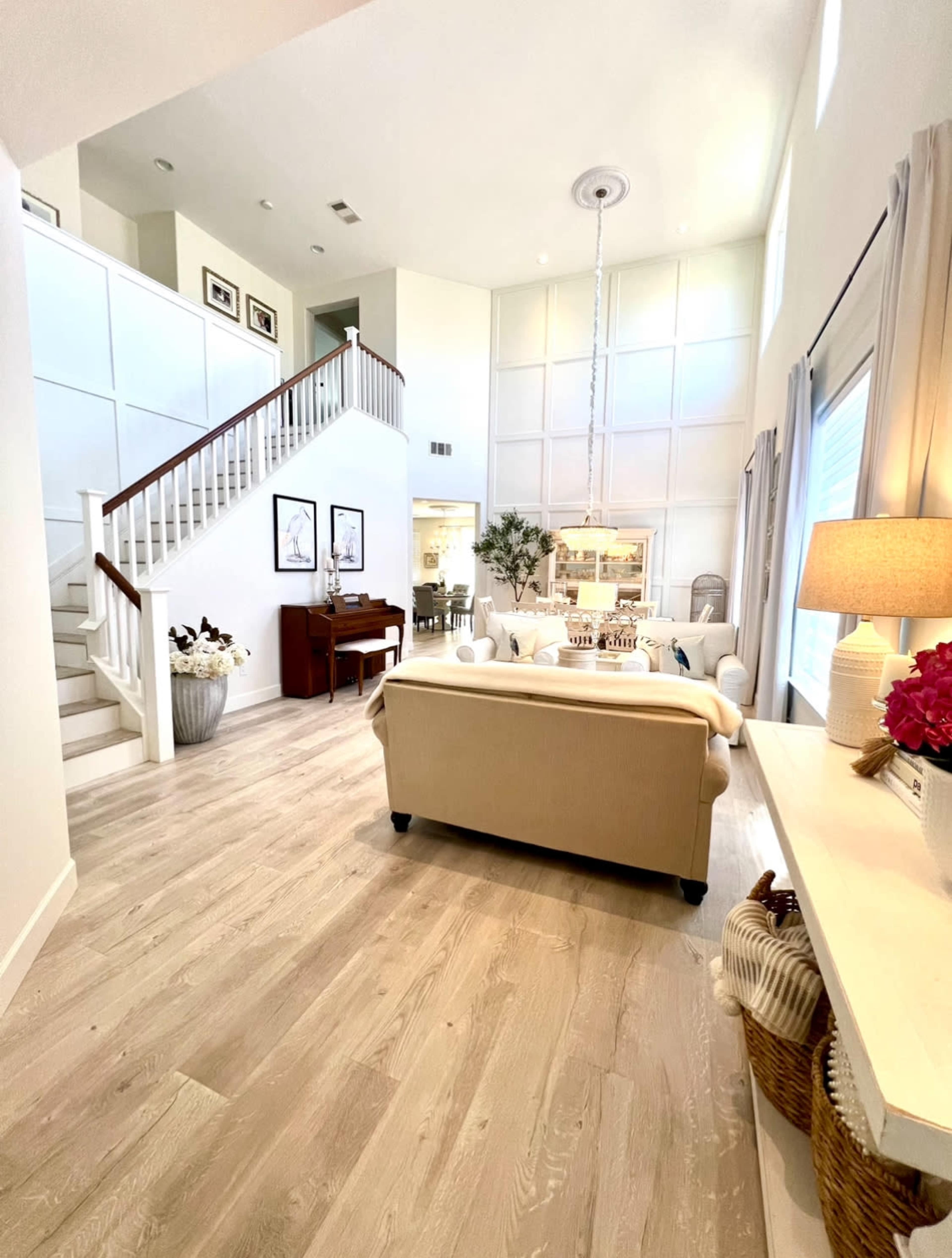 A spacious living room features a light-colored sofa, a piano, and a staircase leading to an upper level, with large windows allowing natural light to fill the space.