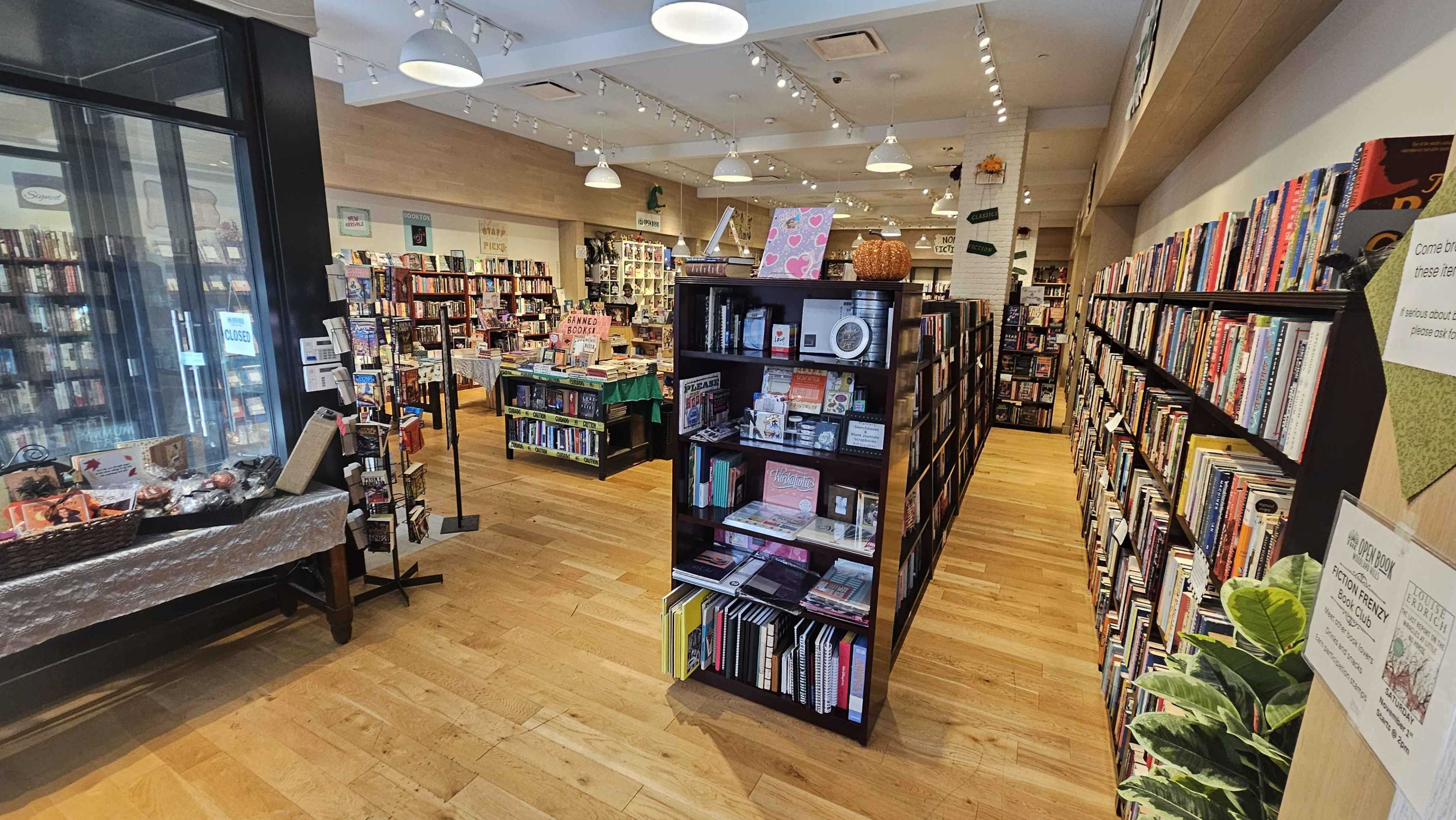 The image shows the interior of a bookstore with wooden flooring, shelves filled with books on either side, and tables displaying merchandise in a well-lit space.