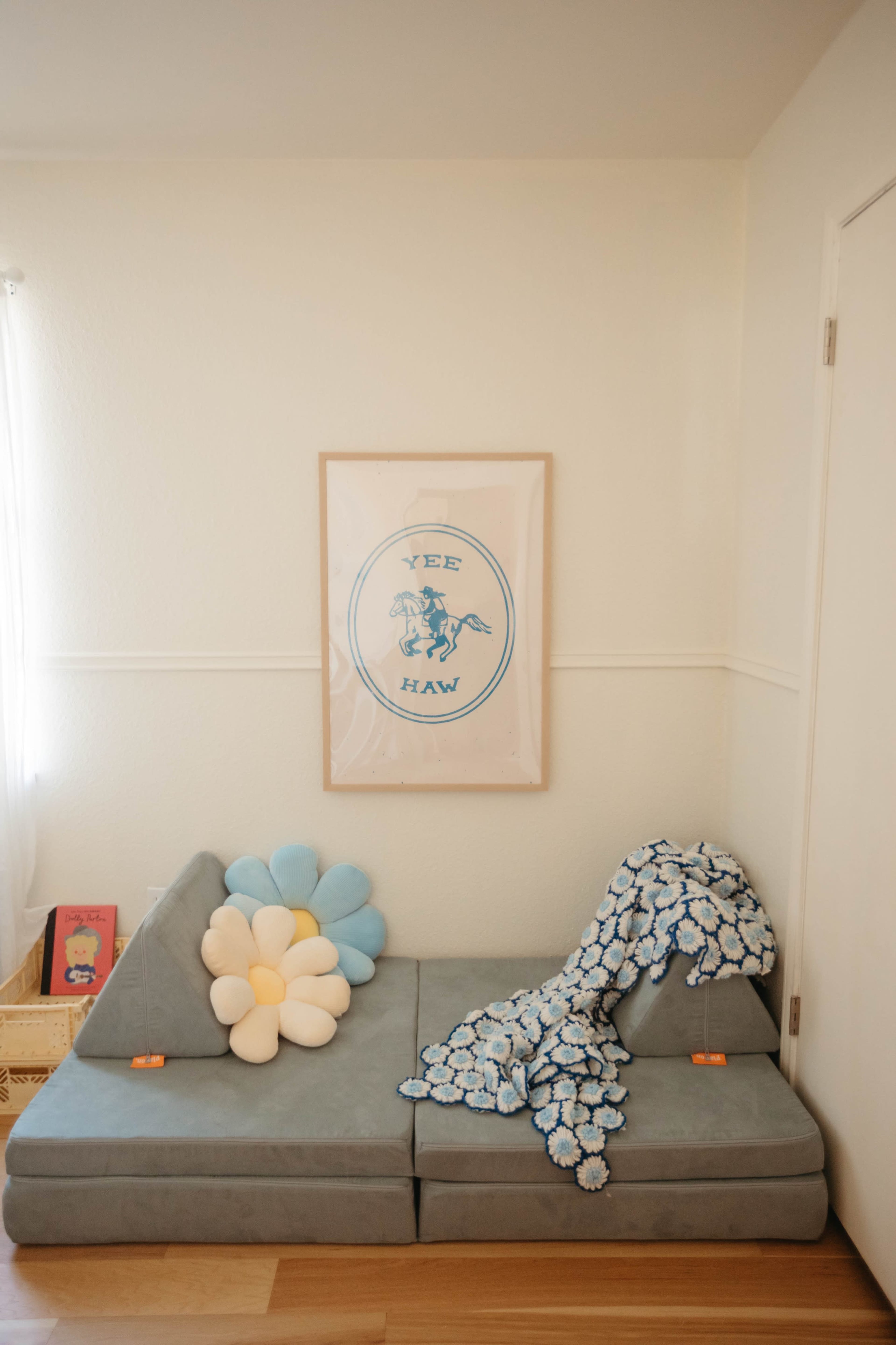 A cozy corner features a light gray sectional sofa adorned with a large flower-shaped pillow and a patterned throw blanket, next to a framed print on the wall.