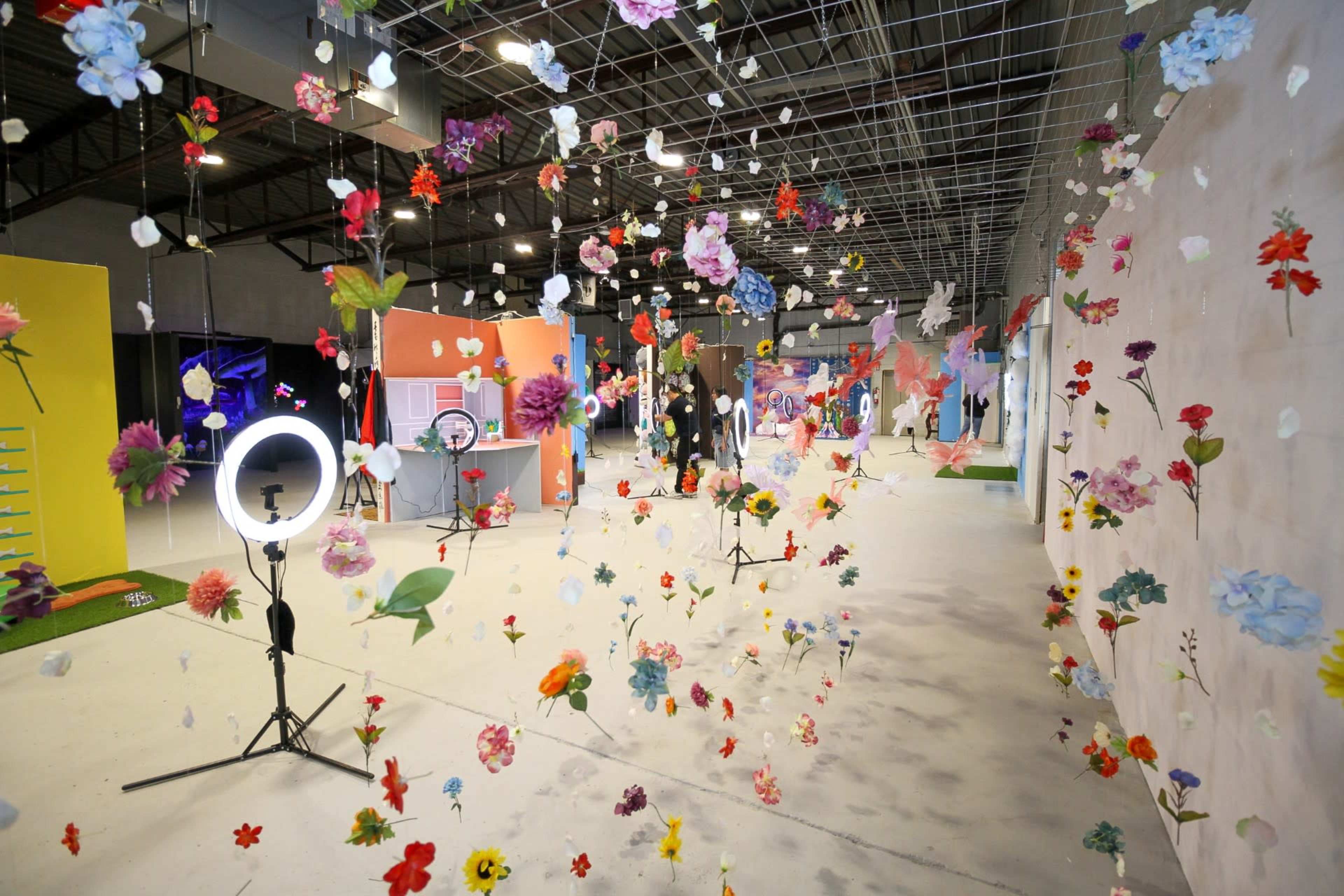 An art installation in a spacious gallery, featuring colorful flowers suspended from above and various colorful backdrops.