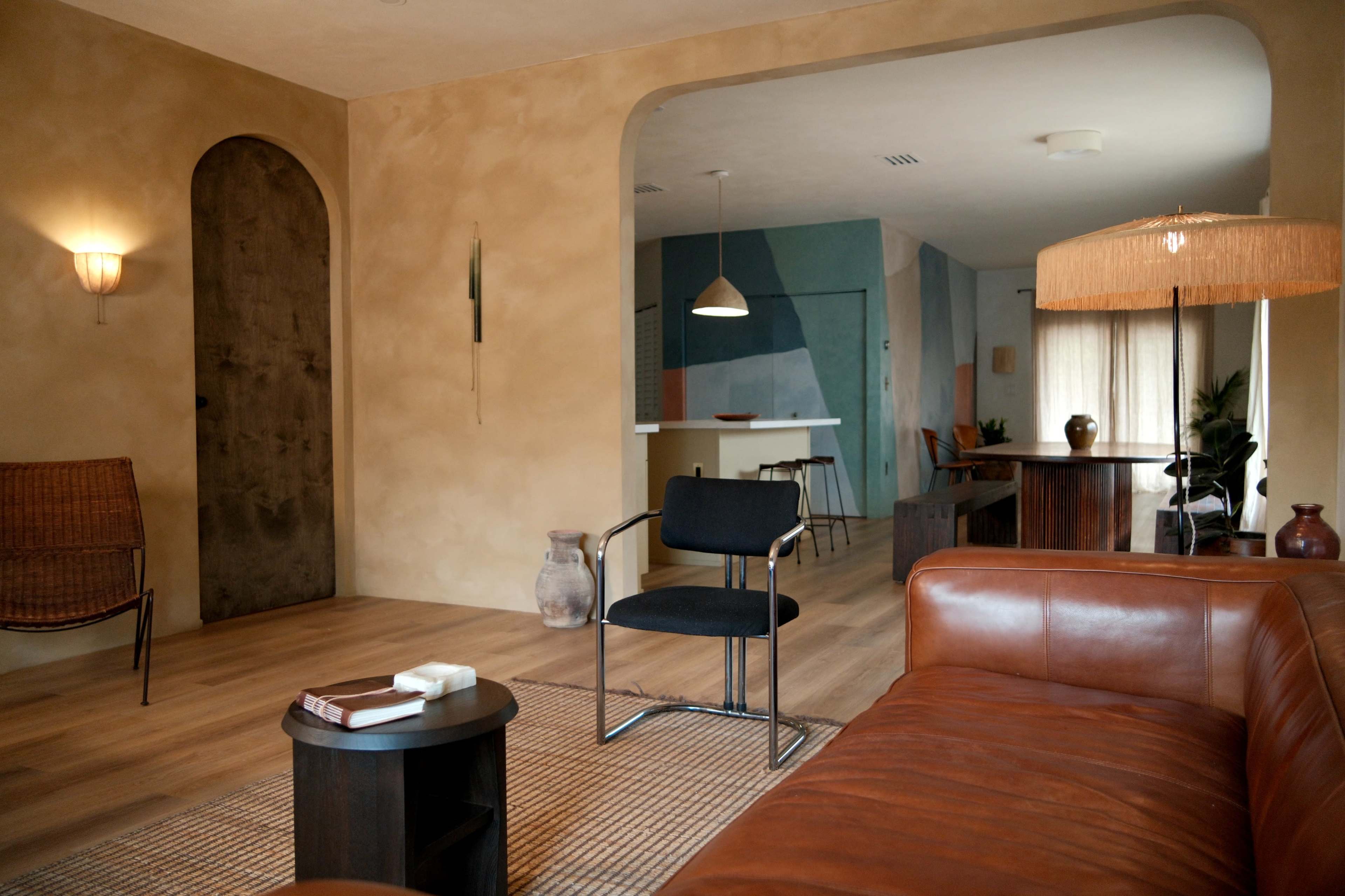 The image depicts a modern living room with a brown leather sofa, a black chair, and an archway leading to a kitchen area.