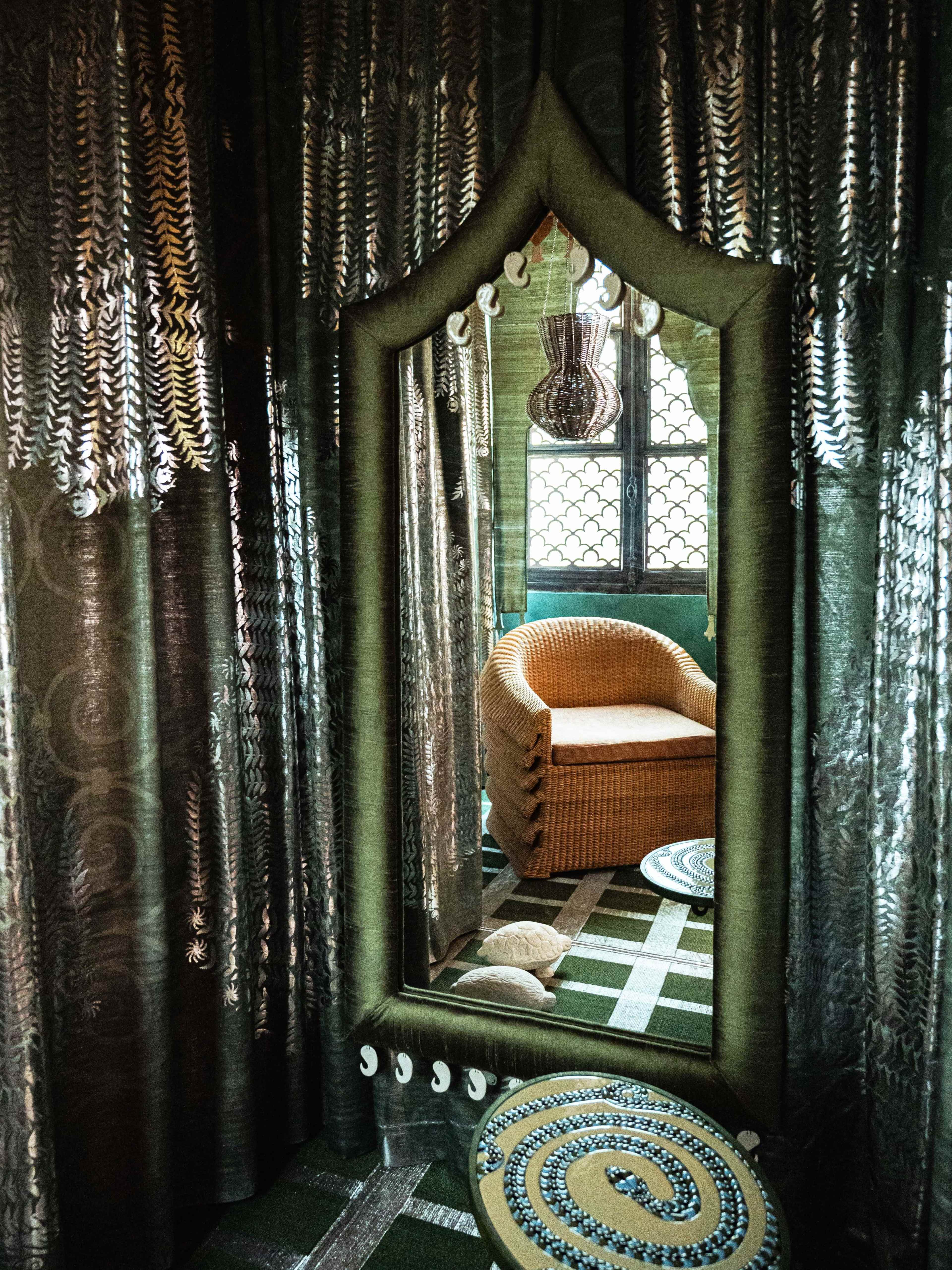 A large, decorative mirror framed with a green fabric reflects a cozy wicker chair and intricately patterned rugs in a richly draped room.