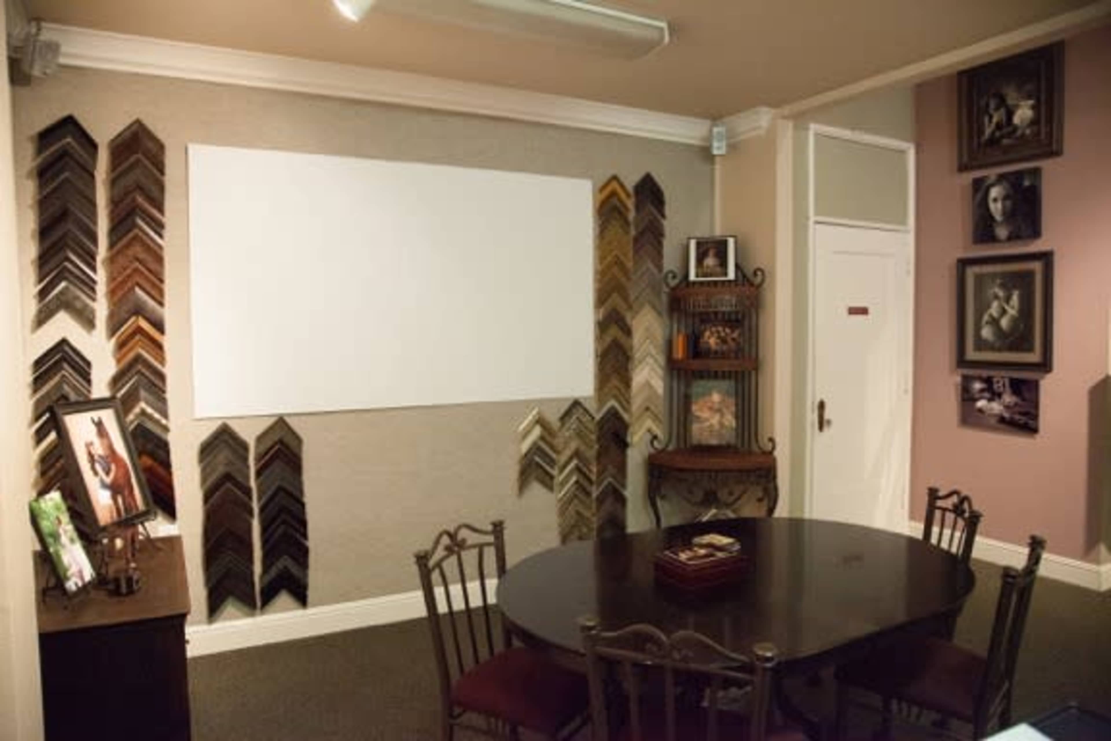 The image shows a room with a round dining table surrounded by chairs, framed artwork on the walls, and various picture frames displayed beside a blank white canvas.