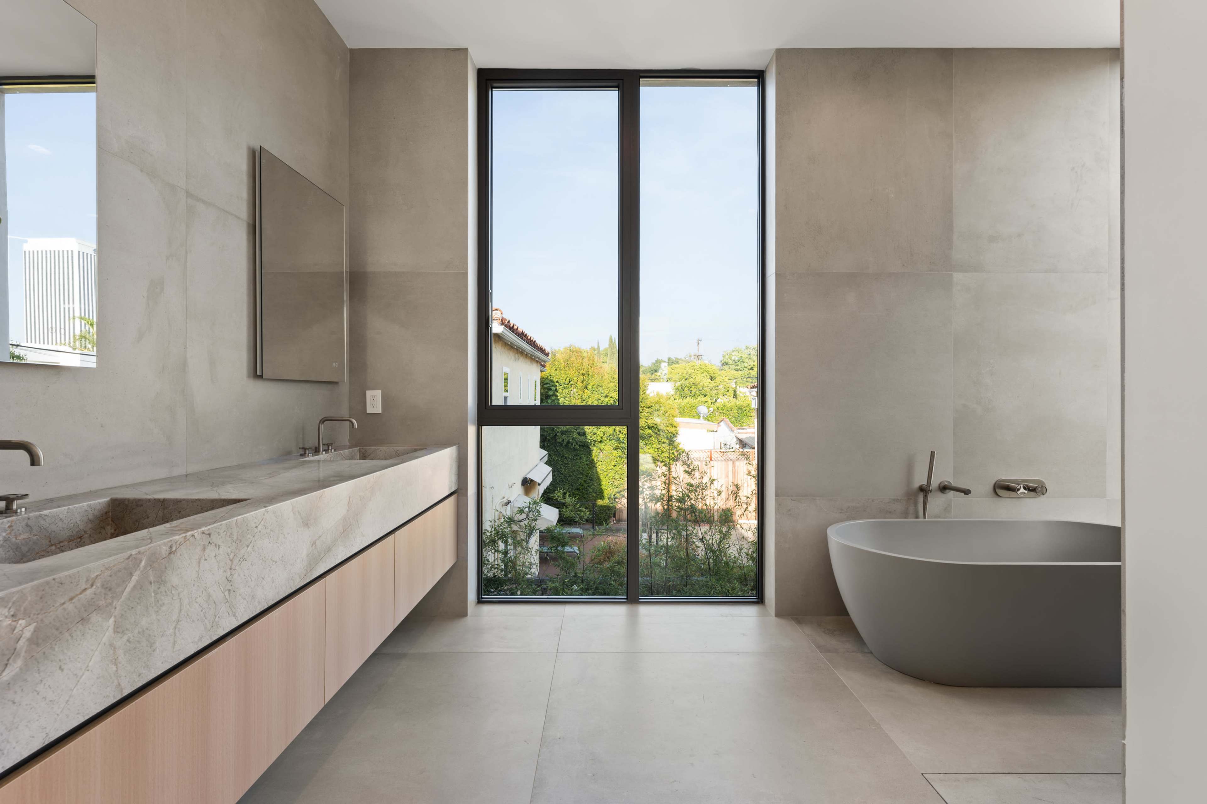 A modern bathroom featuring a dual-sink vanity with a marble countertop, a freestanding bathtub, and large windows that overlook a garden.