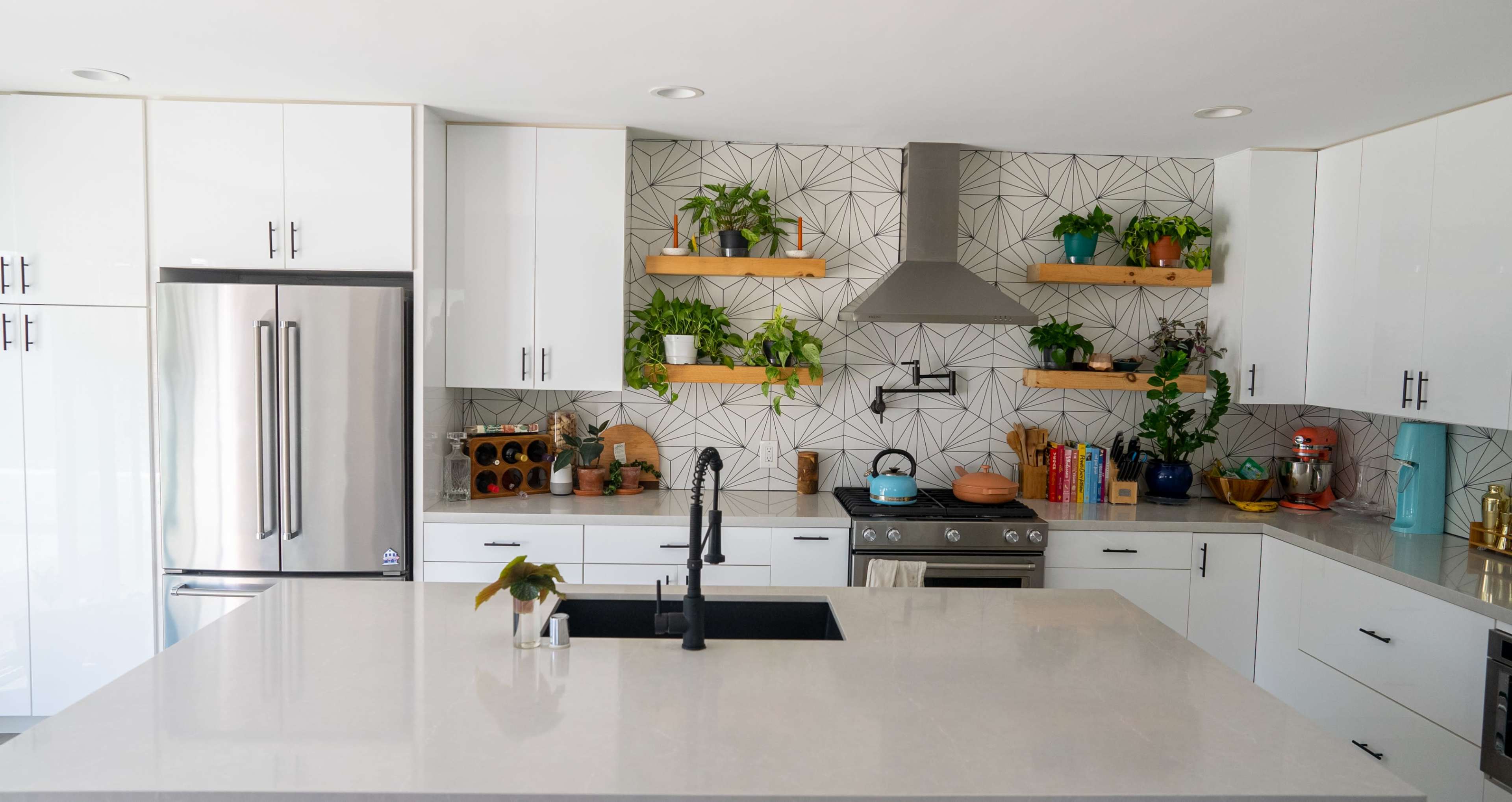 The image shows a modern kitchen with white cabinetry, a large island with a dark sink, and a patterned tile backsplash adorned with greenery on shelves.