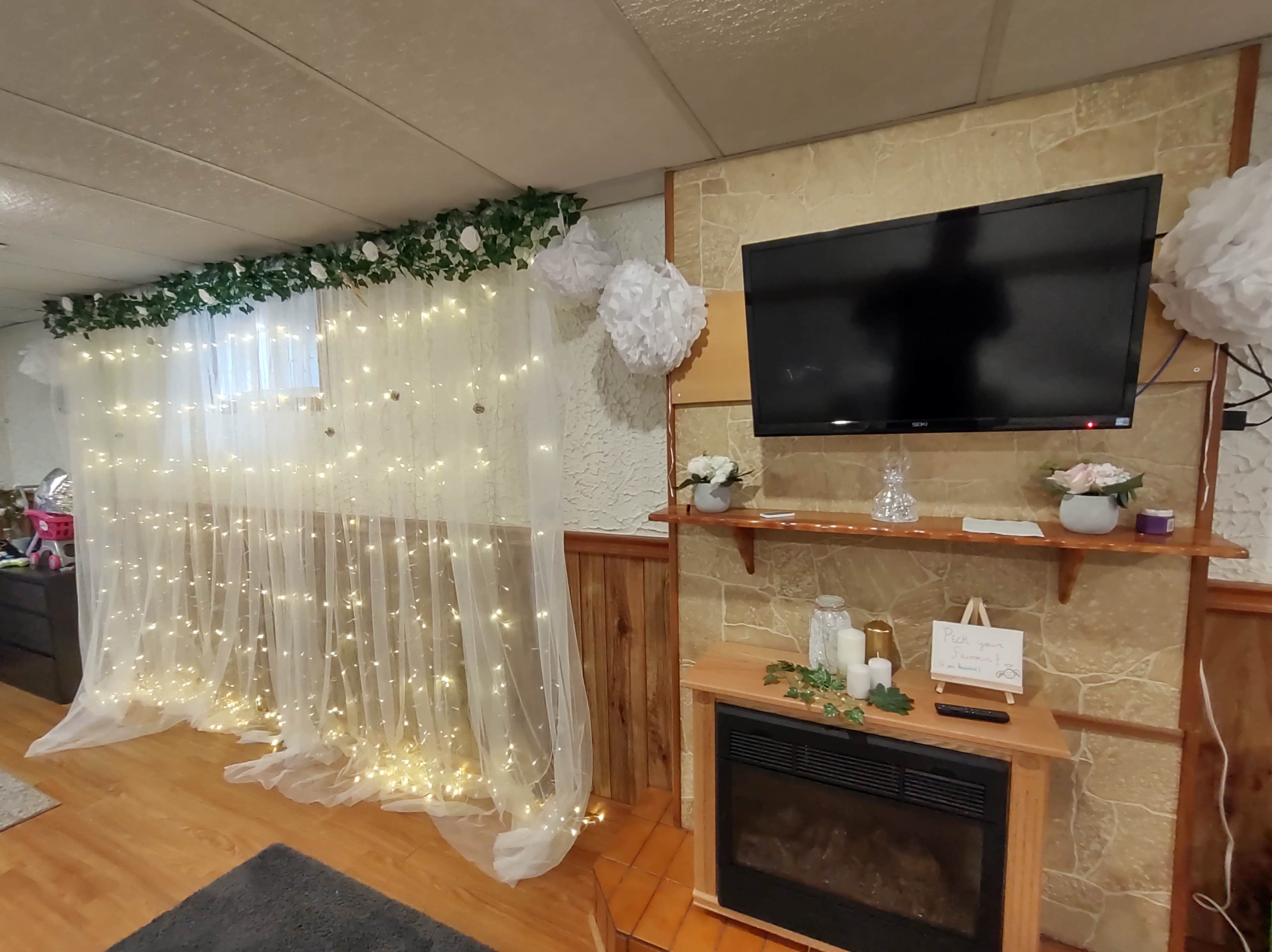 A basement room with a wall draped in sheer curtains adorned with fairy lights, a TV mounted on a stone wall, and a wooden shelf with decorative items above a fireplace.