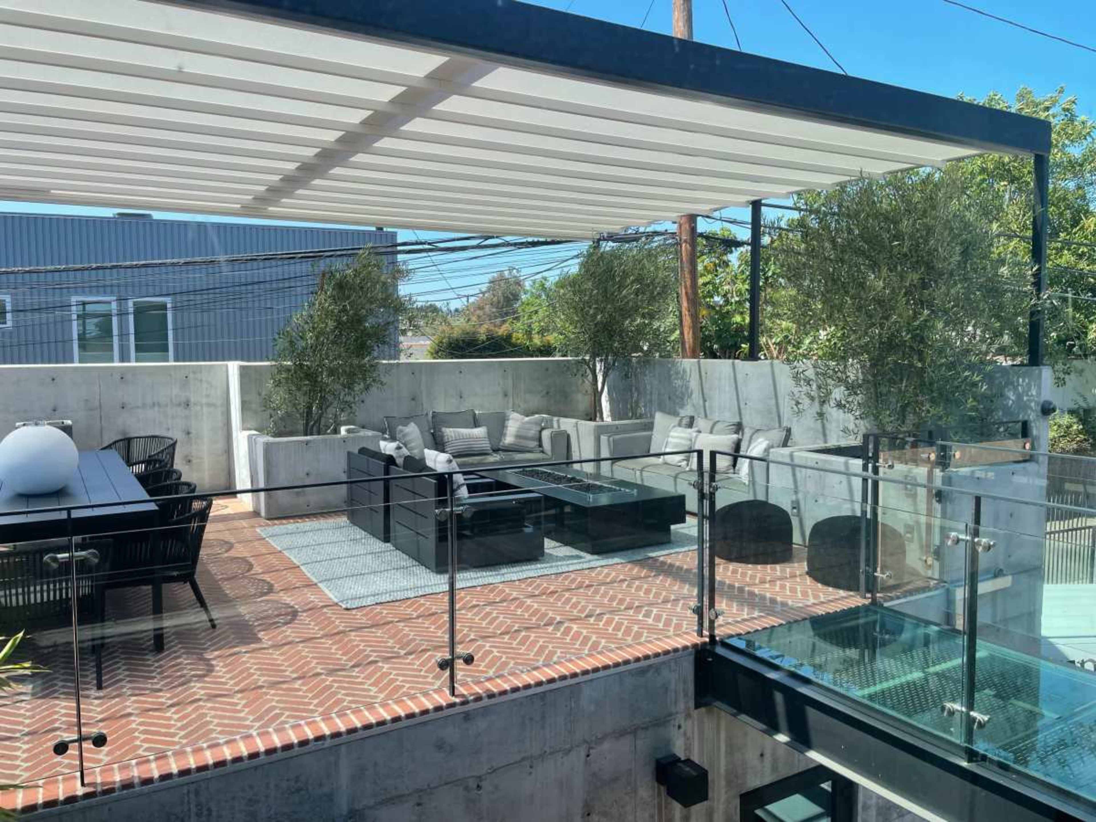 A spacious outdoor patio with sectional seating, a glass railing, and a modern pergola, surrounded by greenery and brick paving.