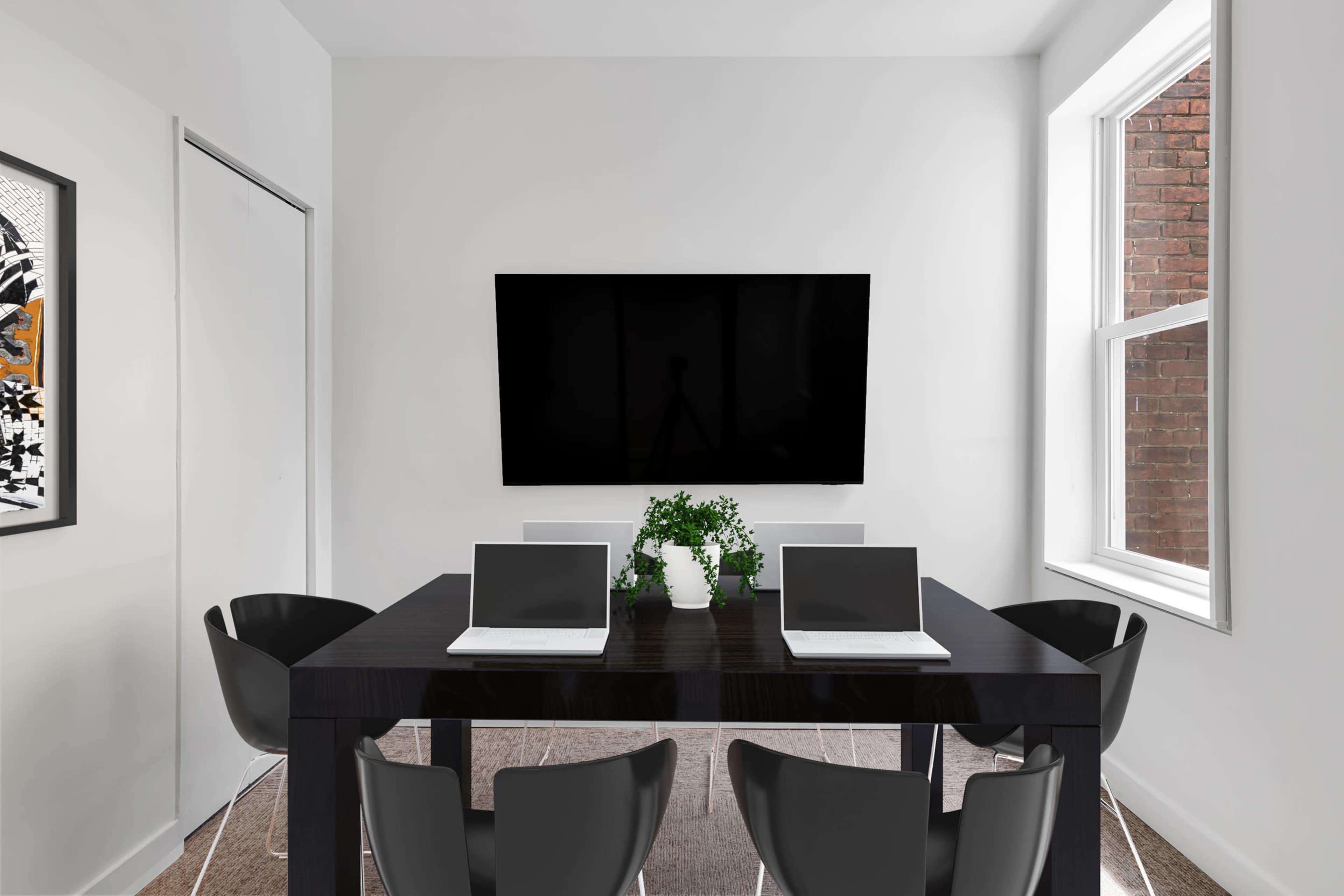 A modern meeting room features a black table with four chairs, two laptops, a green plant centerpiece, and a large television mounted on the white wall.