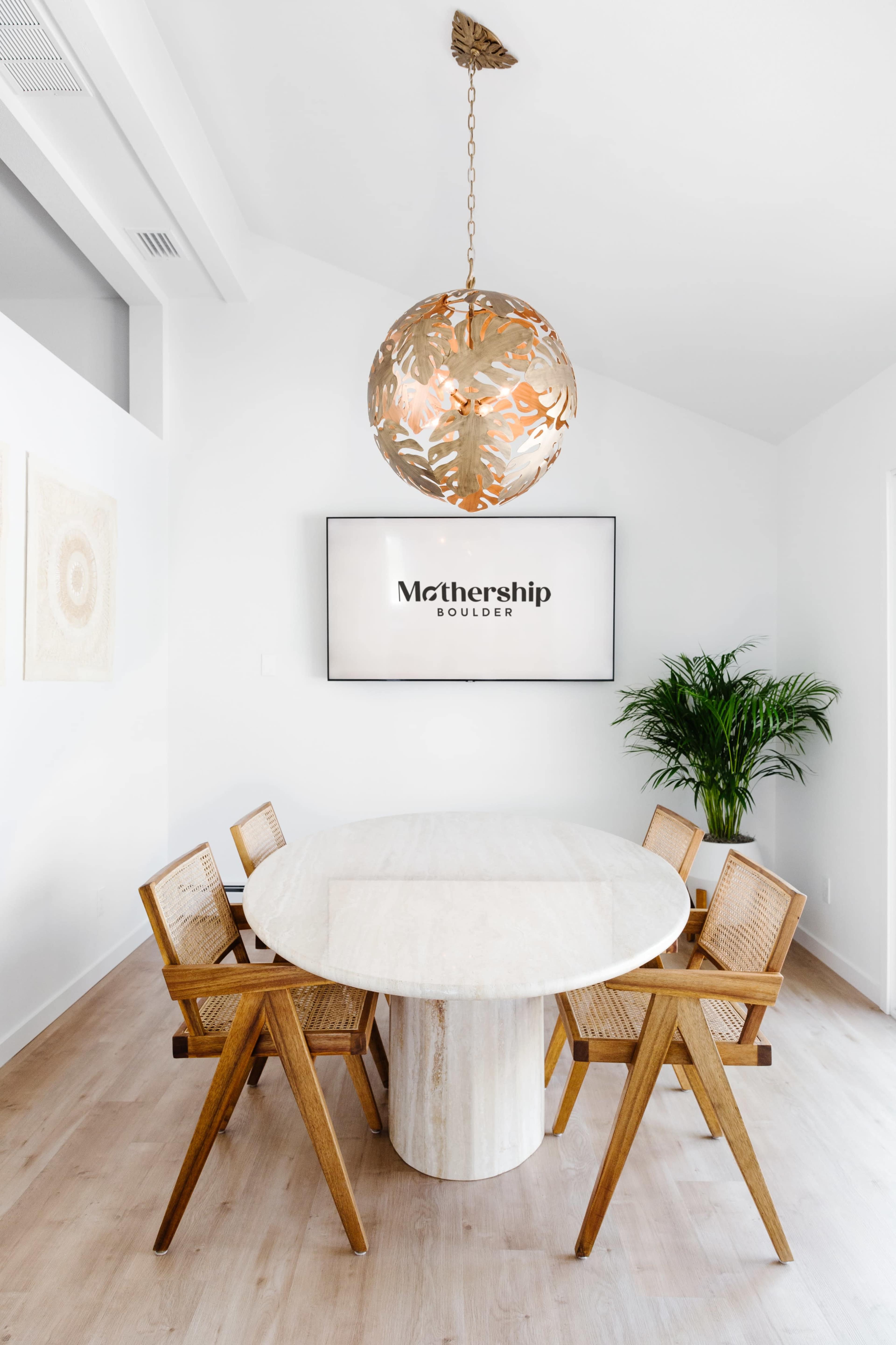 A round dining table with wooden chairs is set in a bright room featuring a television and a hanging pendant light with a floral design.