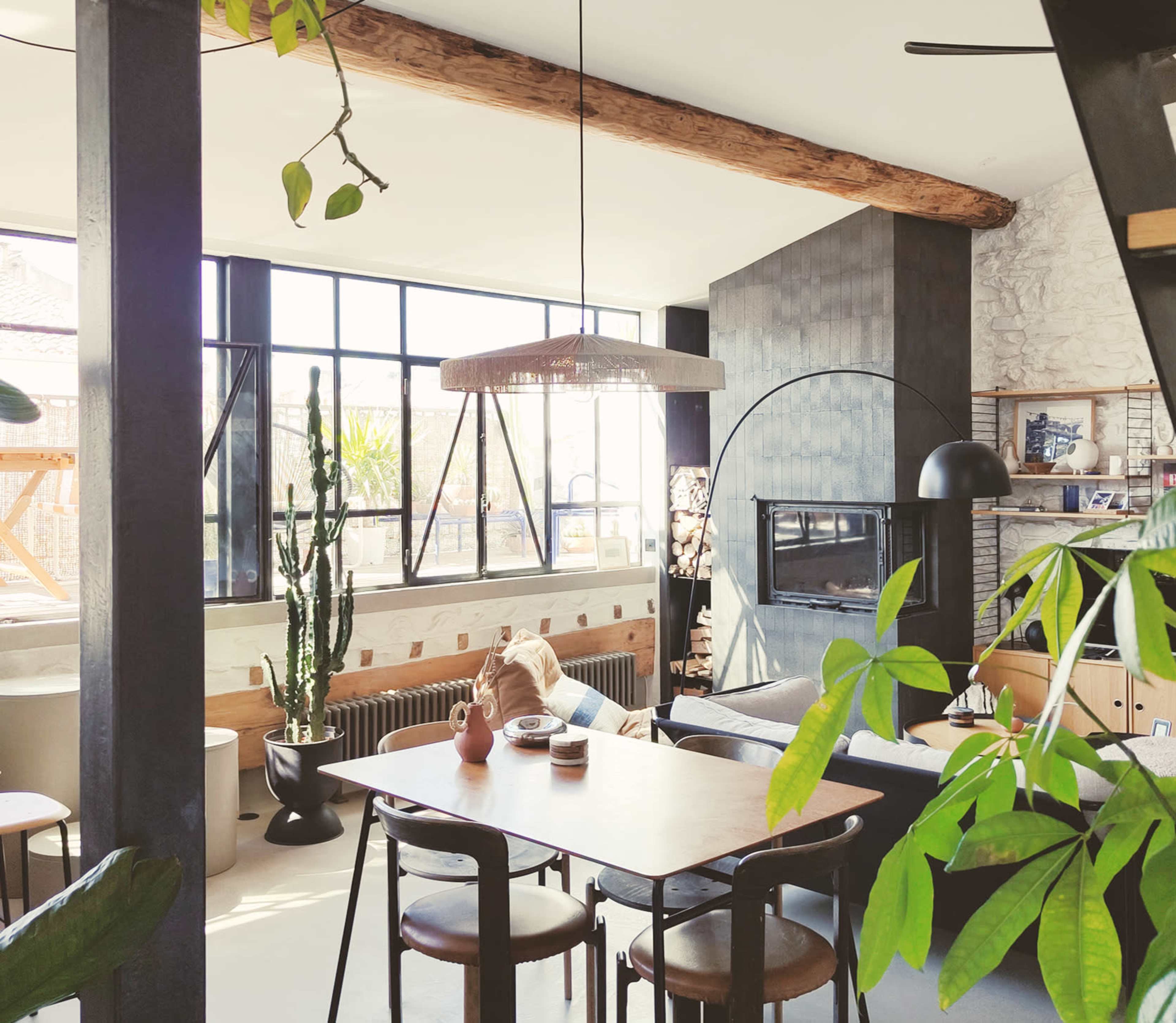 The image shows a sunlit living space featuring a dining table with chairs, large windows, indoor plants, and a fireplace.