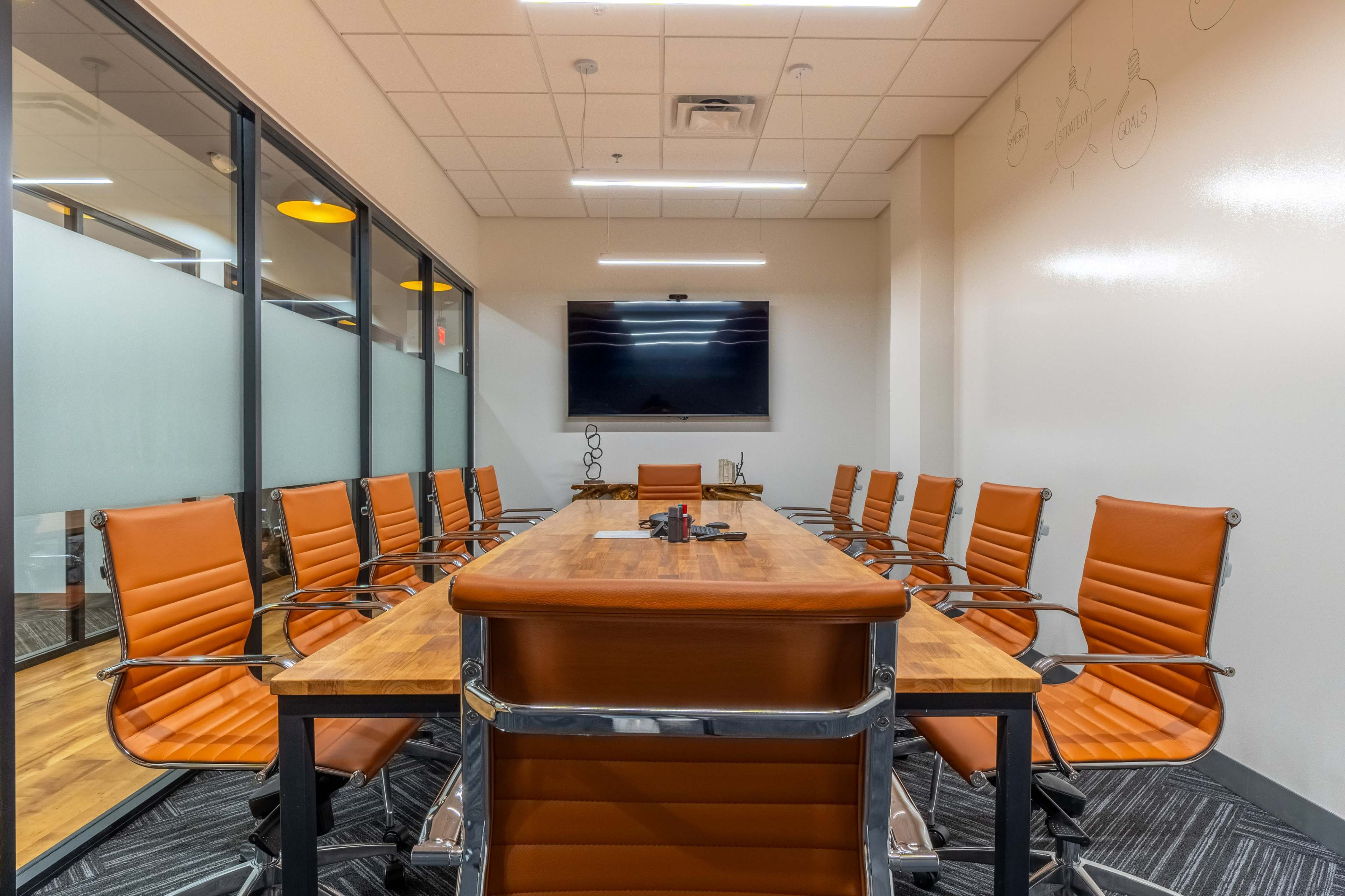 A modern conference room features a long wooden table surrounded by orange chairs and a wall-mounted television.