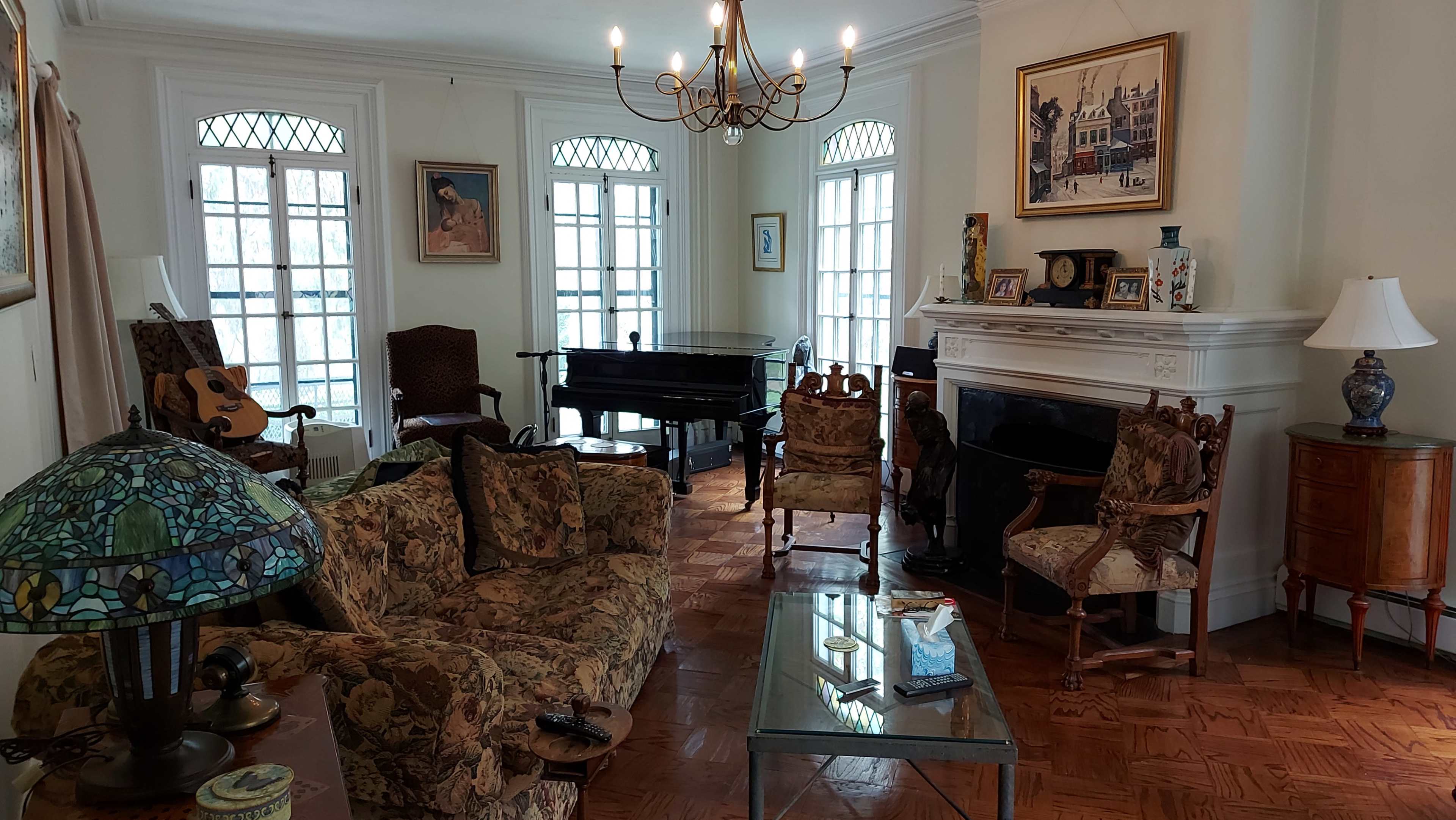 The living room features a grand piano, floral-patterned sofas, and a vintage lamp, all arranged around a central coffee table.