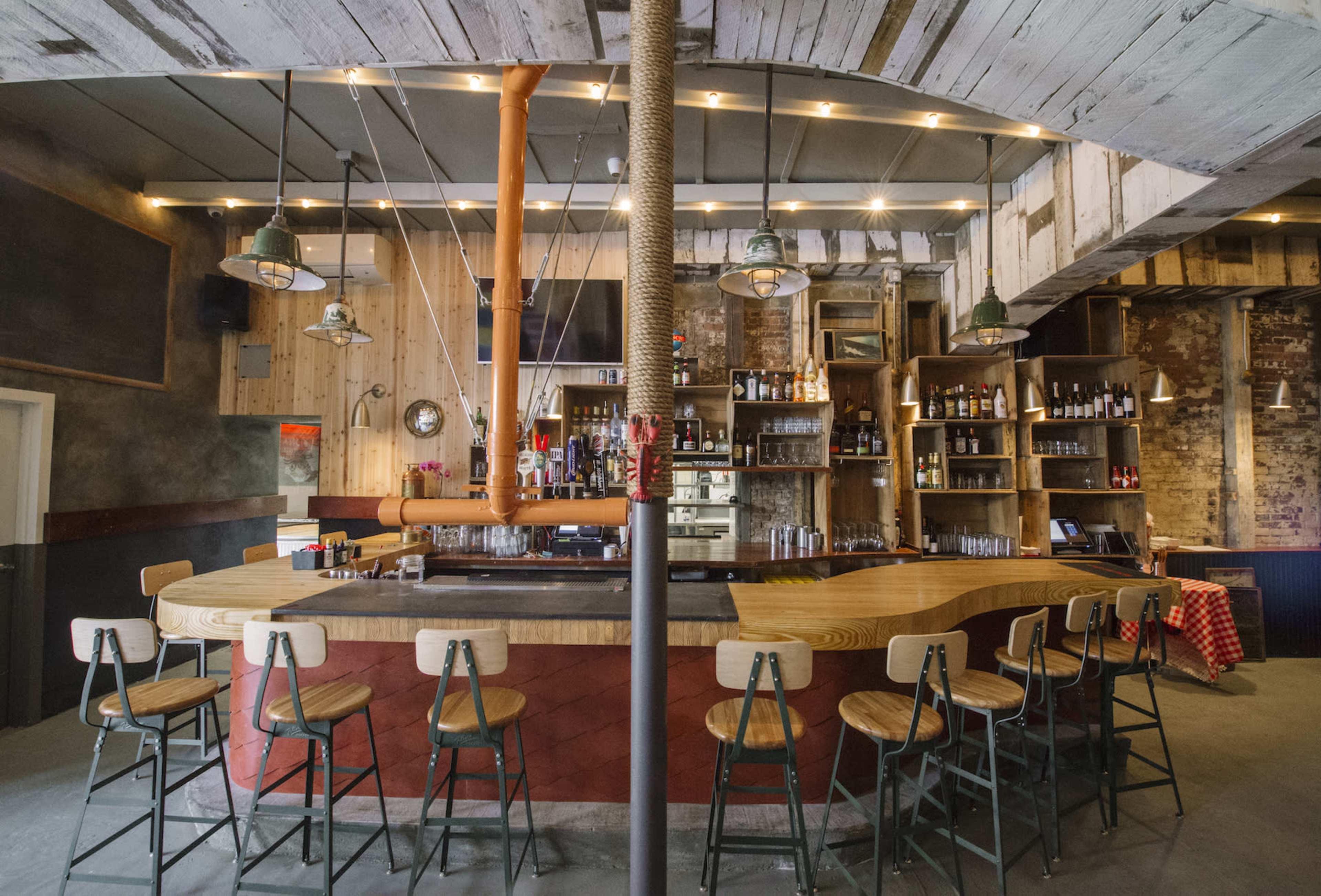 The image shows a rustic bar with wooden stools arranged around a curved counter, illuminated by pendant lights amidst a backdrop of shelves stocked with various bottles.