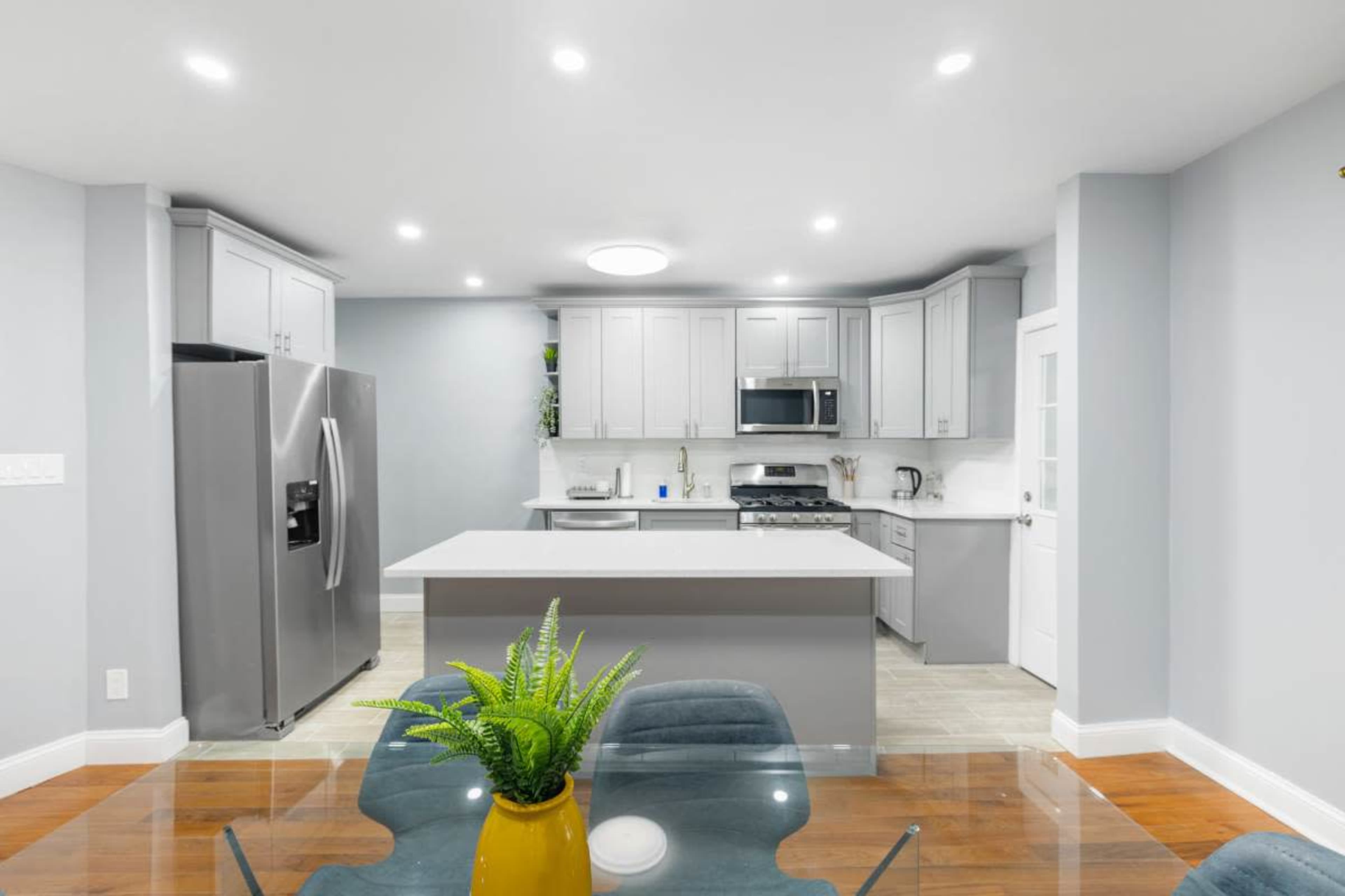 A modern kitchen with gray cabinetry, stainless steel appliances, and a large island, featuring a glass dining table in the foreground.